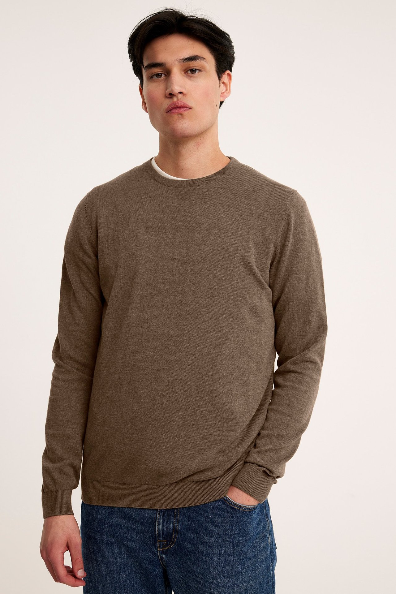 Framsida av en man i en mörkbeige melerad finstickad bomullströja och blå jeans.