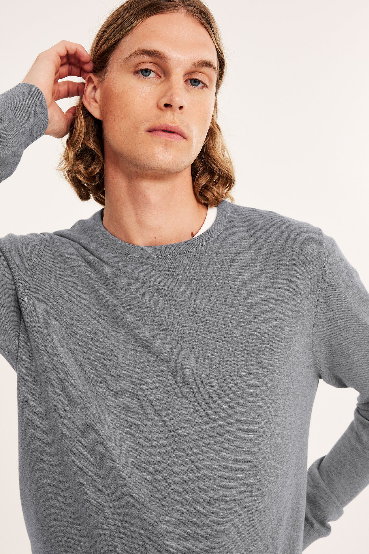 Närbild framifrån: man bär en ljusgrå melerad finstickad bomullströja över en vit t-shirt.