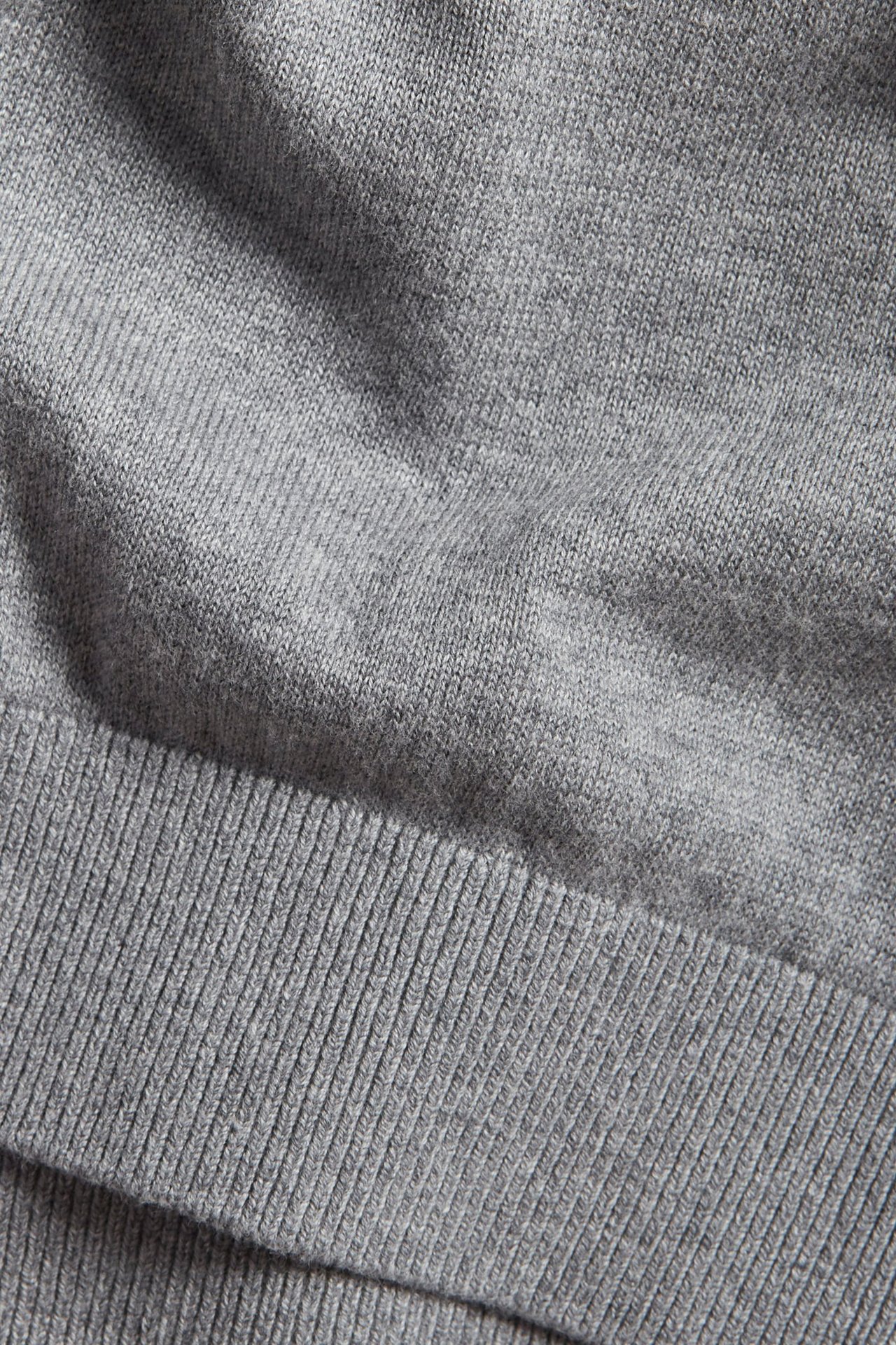 Zbliżenie na jasnoszary melanżowy, drobno dziergany sweter bawełniany z prążkowanym dołem.