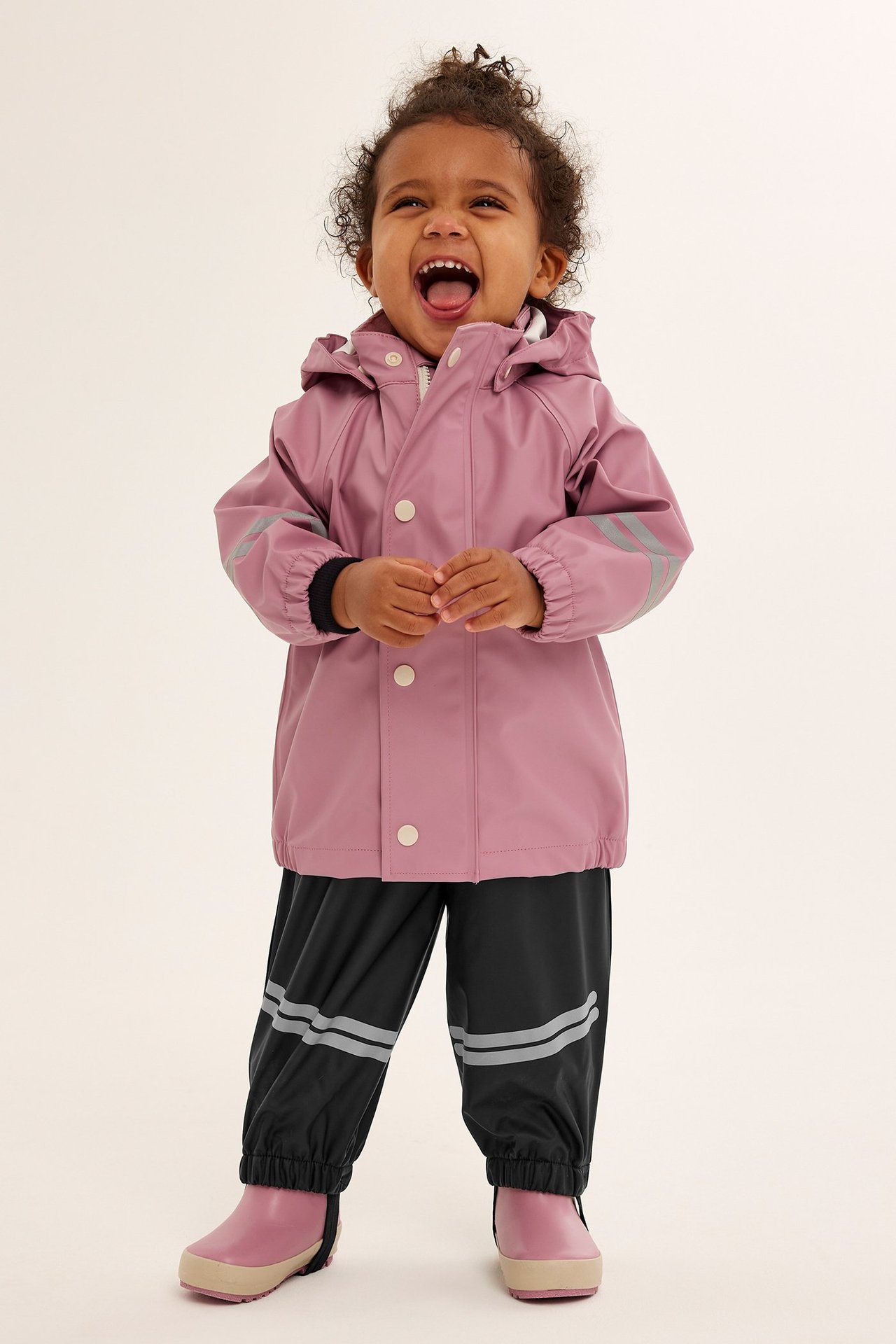 Forfra, en smilende baby i svarte regnbukser med refleksstriper, en rosa regnjakke og rosa regnstøvler.