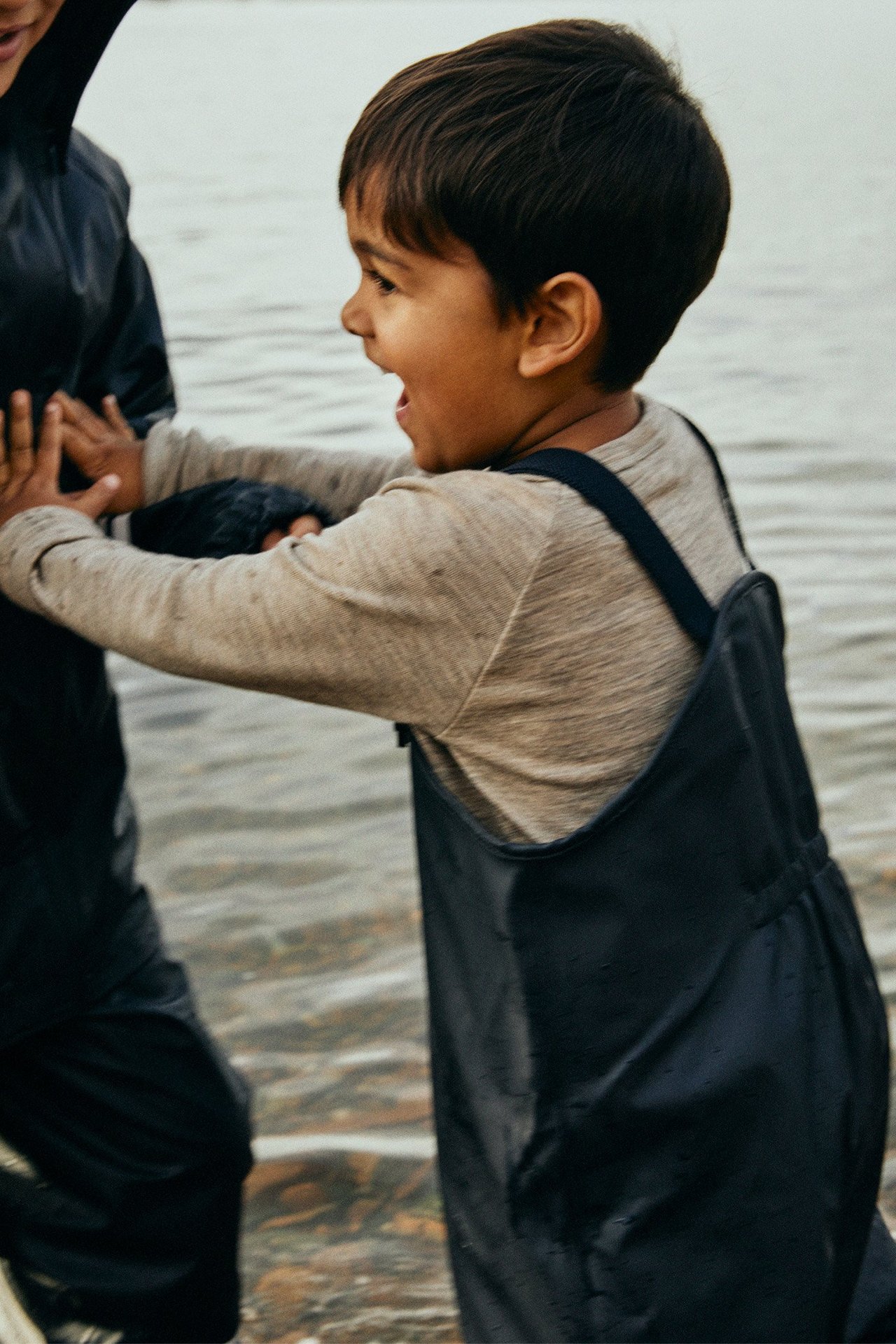 Sidovy av ett leende barn i mörkblå fleecefodrade regnbyxor med hängslen över en ljusbrun långärmad t-shirt.