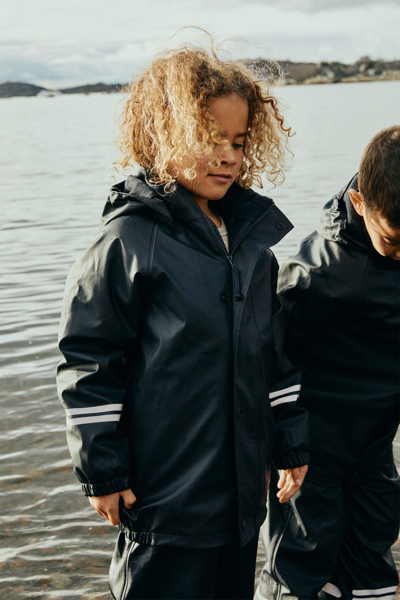 Widok z przodu: Dziecko w ciemnoniebieskiej kurtce przeciwdeszczowej z polarem, odblaskami, zamkiem/guzikami i pasującymi spodniami.