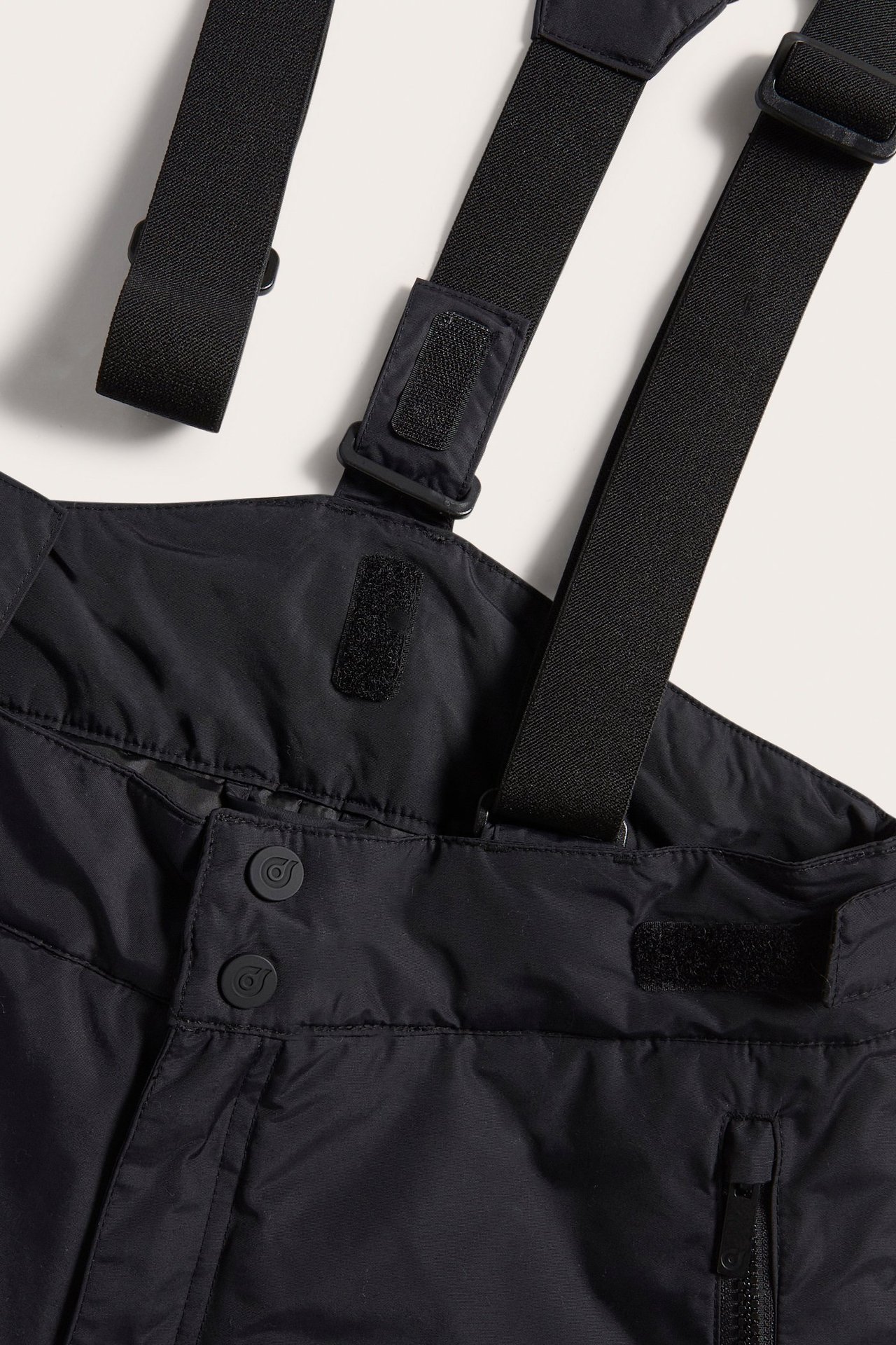 Zbliżenie na czarne spodnie narciarskie z szelkami, pokazujące guziki, rzepy i kieszeń na zamek.