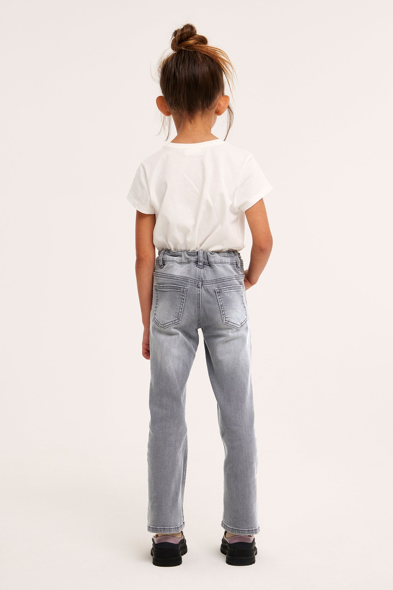 Takaa: Lapsi harmaissa bootcut-farkuissa, joissa painonappi, ja valkoisessa t-paidassa.