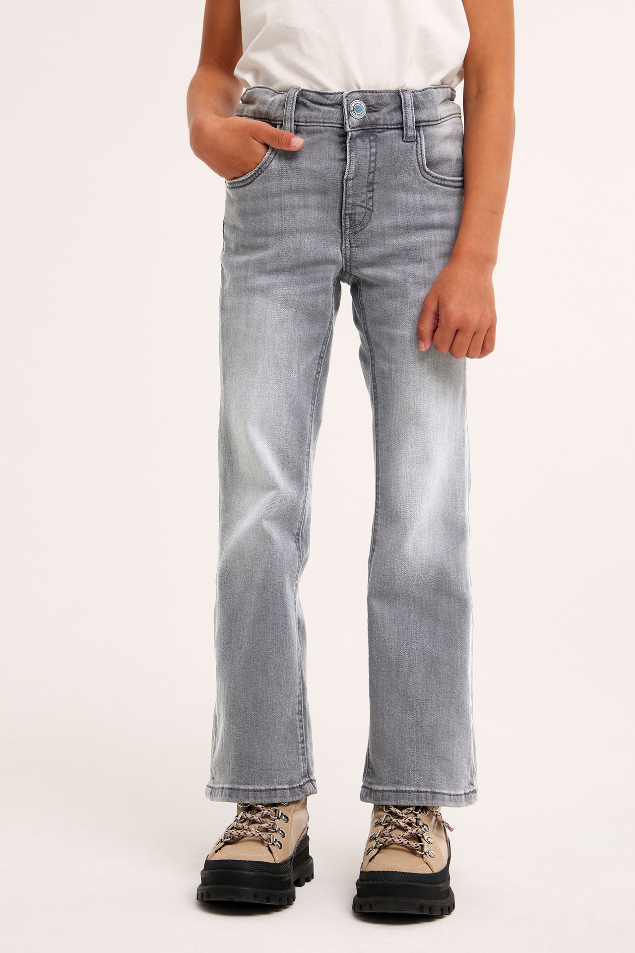 Lapsi haalistuneissa harmaissa bootcut-farkuissa napilla, valkoisessa t-paidassa ja beigenvärisissä paksupohjaisissa kengissä, edestäpäin.
