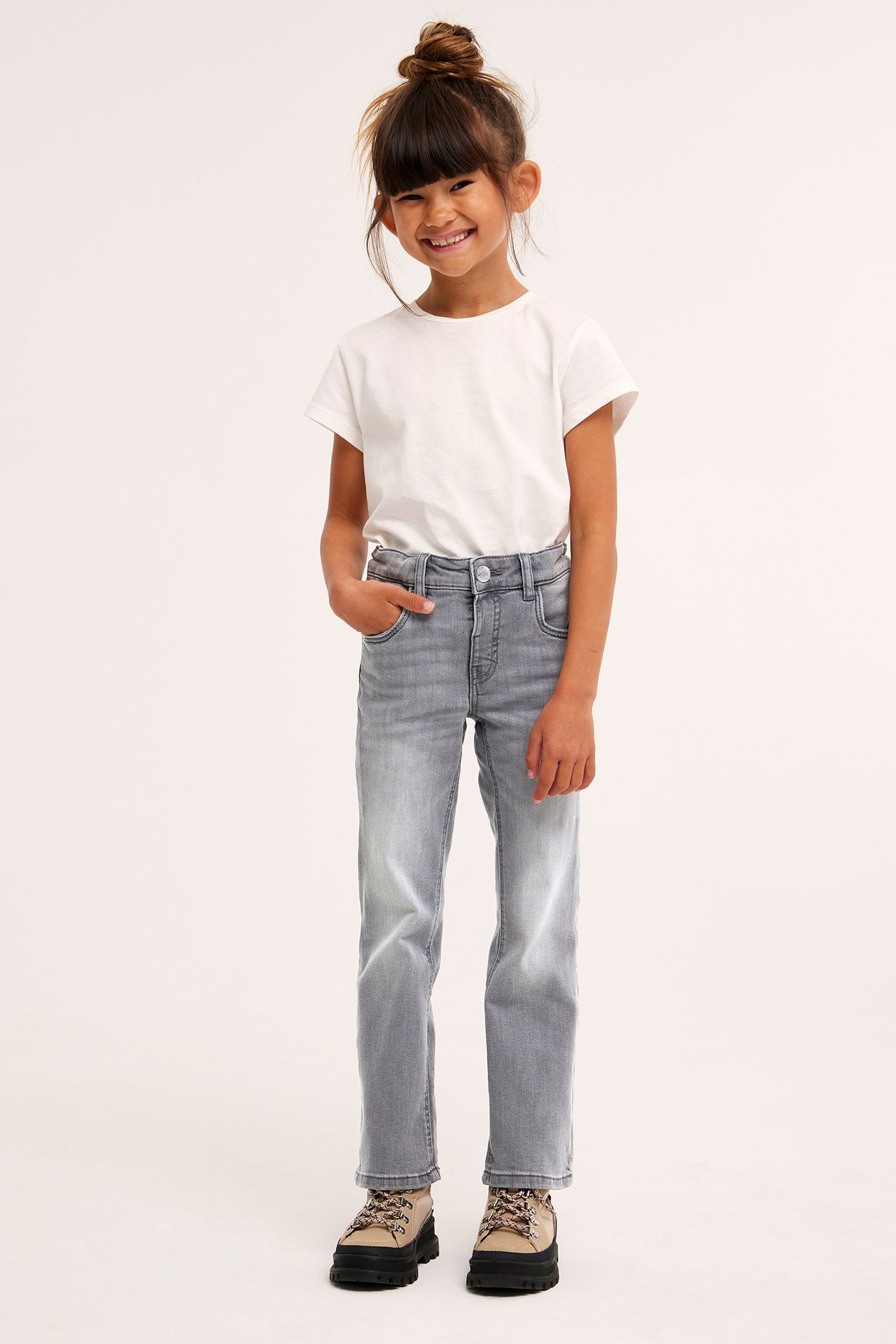 Edestäpäin: lapsi harmaissa denim bootcut-farkuissa painonapilla, valkoisessa t-paidassa ja beigenvärisissä kengissä.