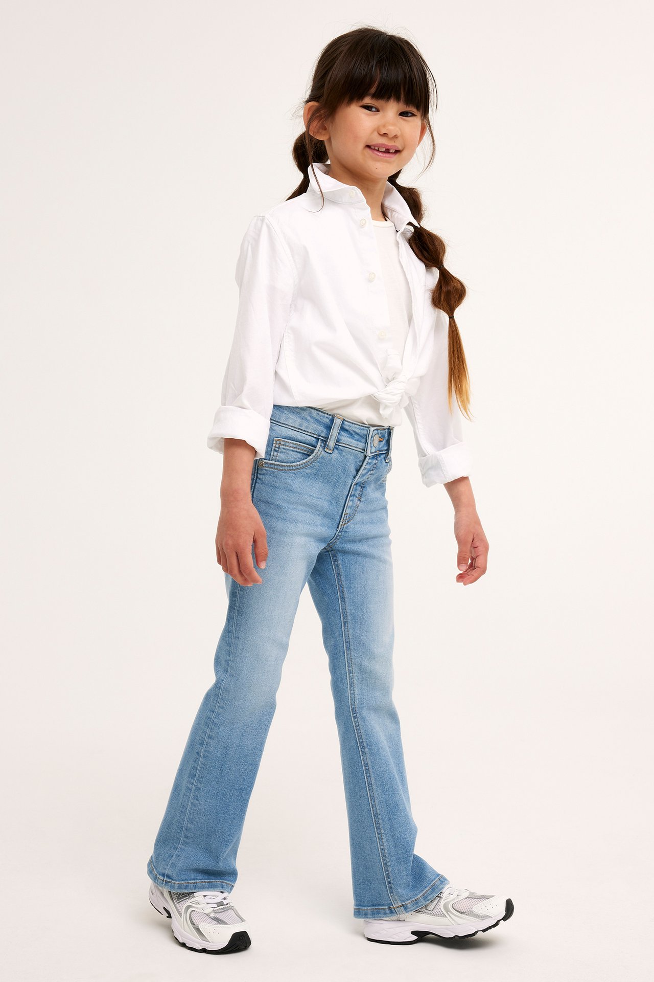 Frontvy av ett barn i ljusblå bootcut-jeans med tryckknapp, en vit skjorta och vita sneakers.