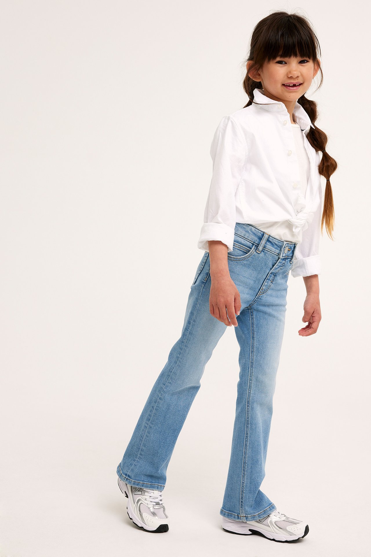 Frontvy av ett barn i ljusblå bootcut-jeans med tryckknapp, en vit skjorta och vita sneakers.