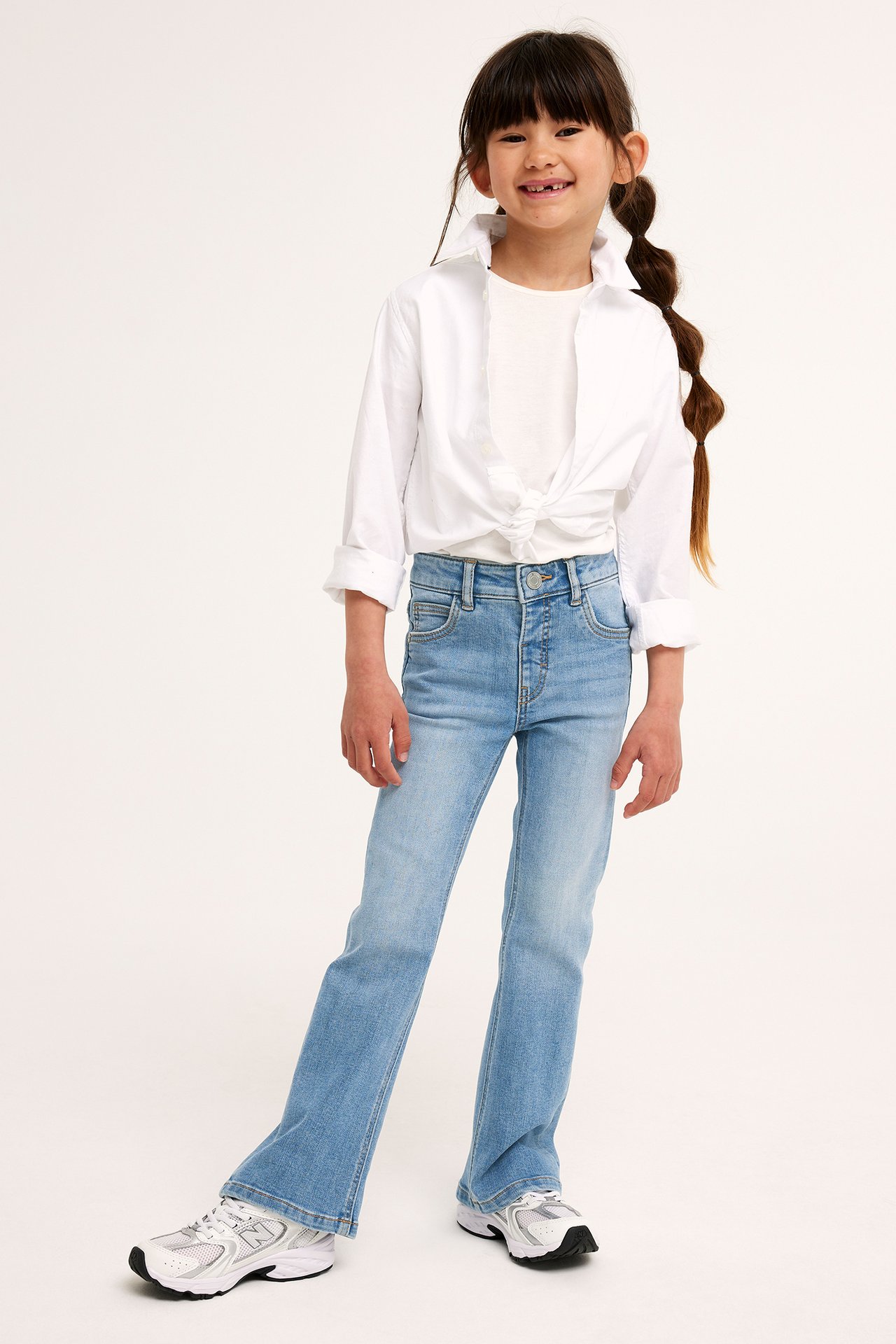 Framifrån: Barn i ljusblå bootcut-jeans, vit t-shirt, knuten vit skjorta och vita sneakers.