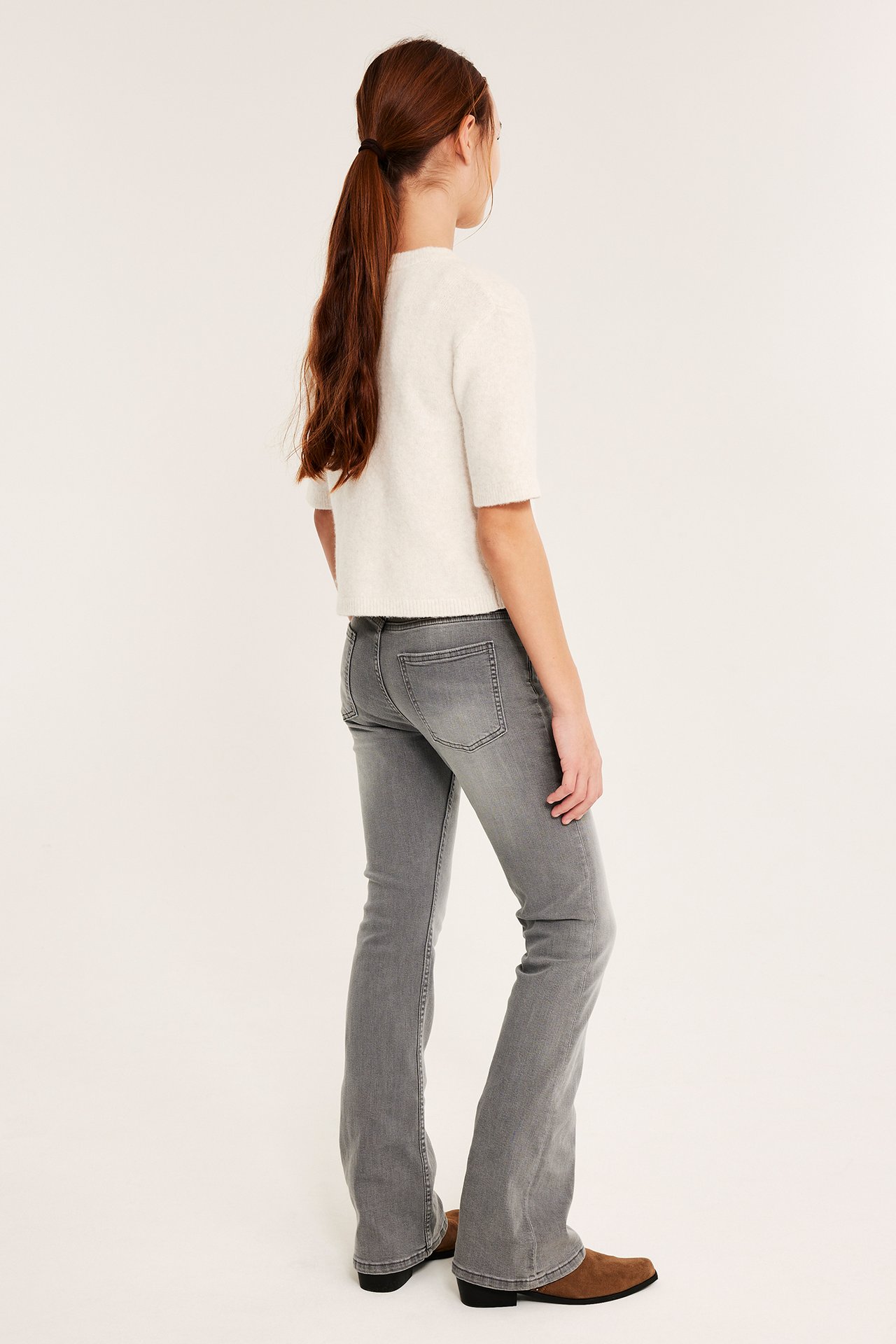 Sidovy av barn i gråa bootcut jeans, vit kortärmad topp och bruna stövlar.