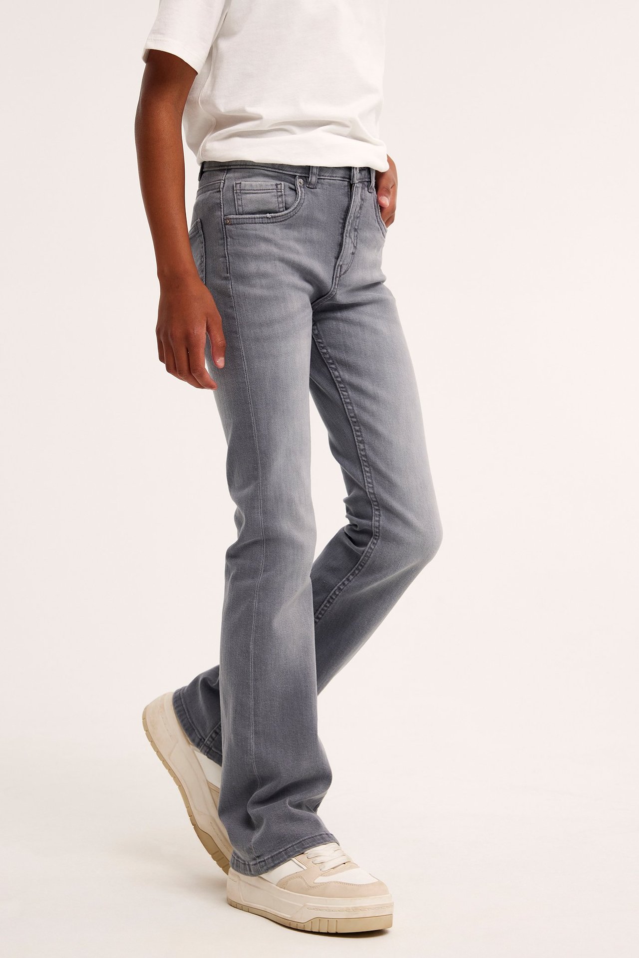 Sivukuva lapsesta harmaissa denim bootcut-farkuissa, valkoisessa t-paidassa ja beigenvärisissä lenkkareissa.