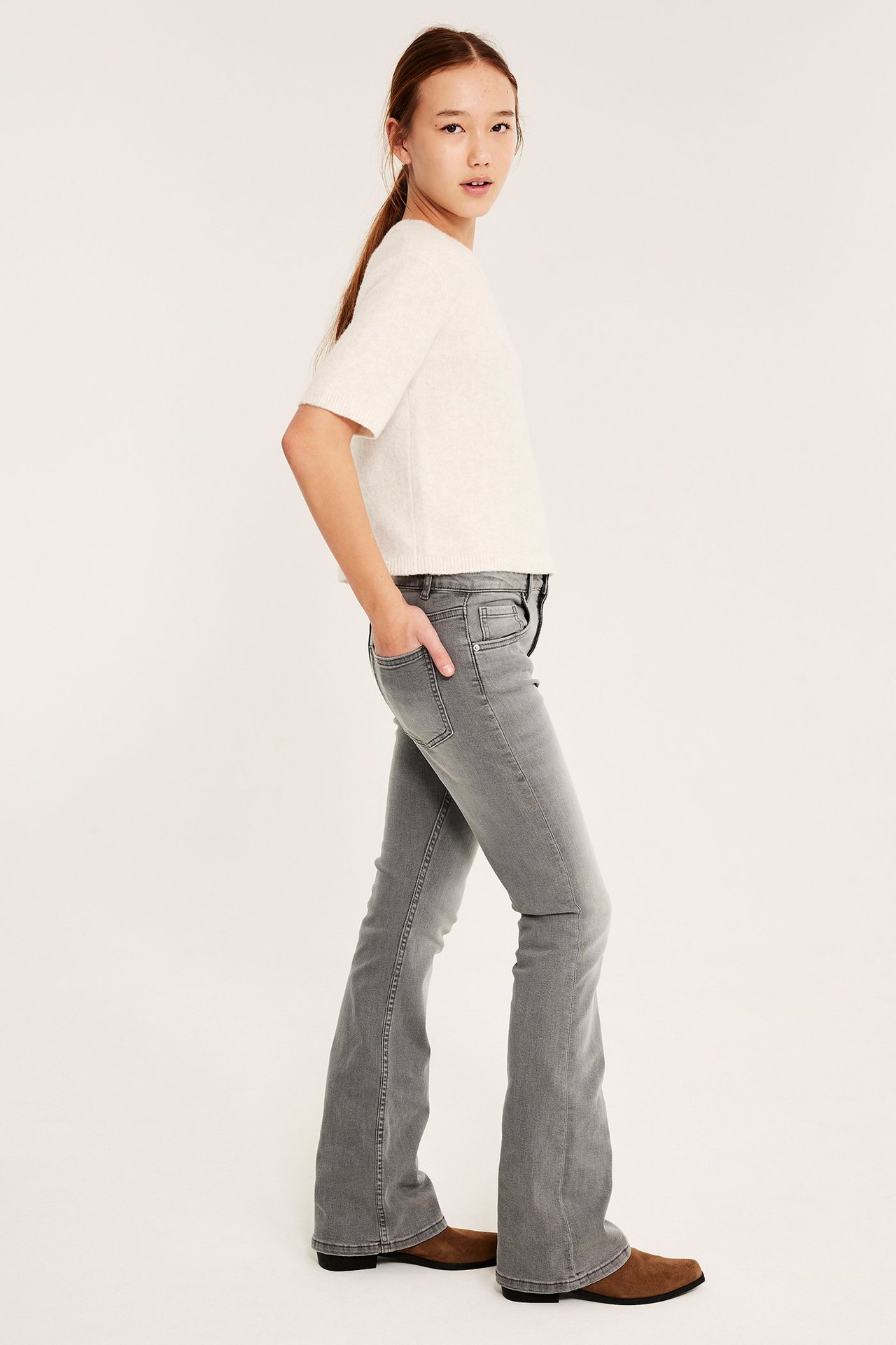 Sivukuva lapsesta harmaissa mid-waist bootcut-farkuissa, kermanvalkoisessa lyhythihaisessa paidassa ja ruskeissa kengissä.