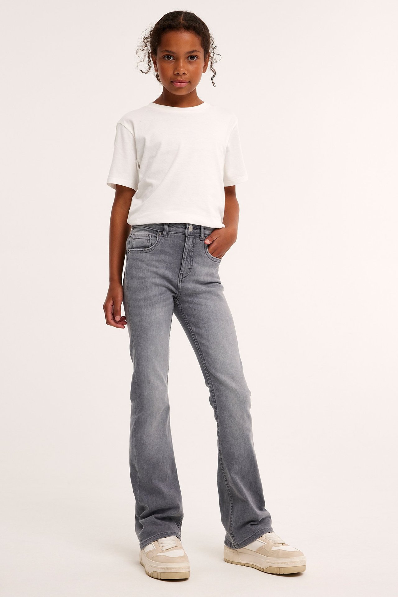 Edestäpäin: lapsi harmaissa bootcut-farkuissa, valkoisessa t-paidassa ja beigenvärisissä lenkkareissa.