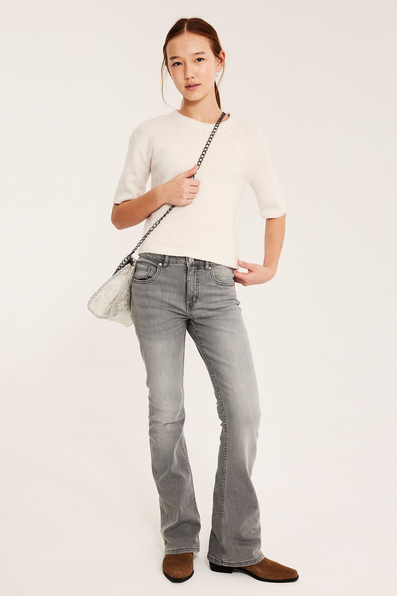 Widok z przodu: dziecko w szarych dżinsach bootcut, kremowym swetrze, brązowych butach i błyszczącej torebce.