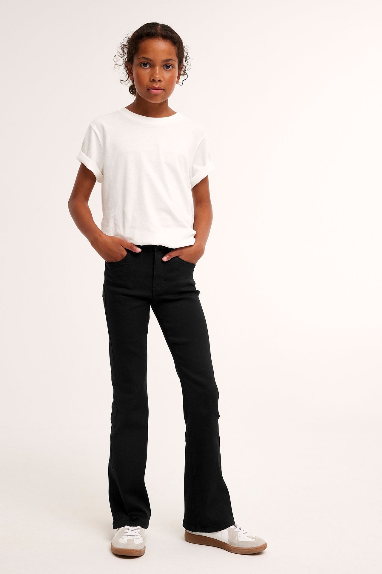 Edestäpäin, lapsi mustissa bootcut-farkuissa, valkoisessa t-paidassa ja valkoisissa tennareissa.