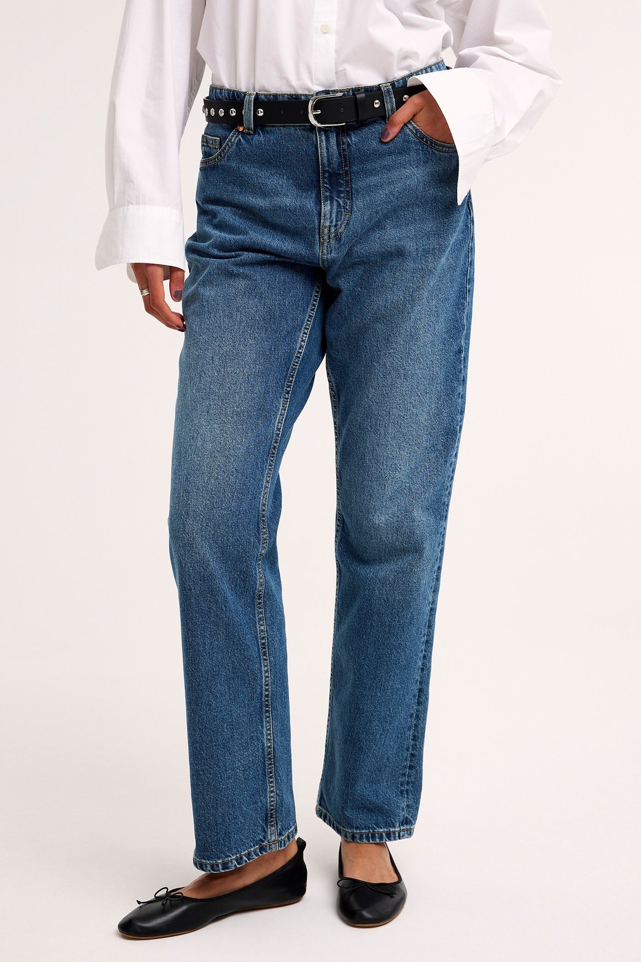 Framifrån, kvinna i blå raka jeans med hög midja, vit skjorta, svart nitbälte och svarta ballerinaskor.