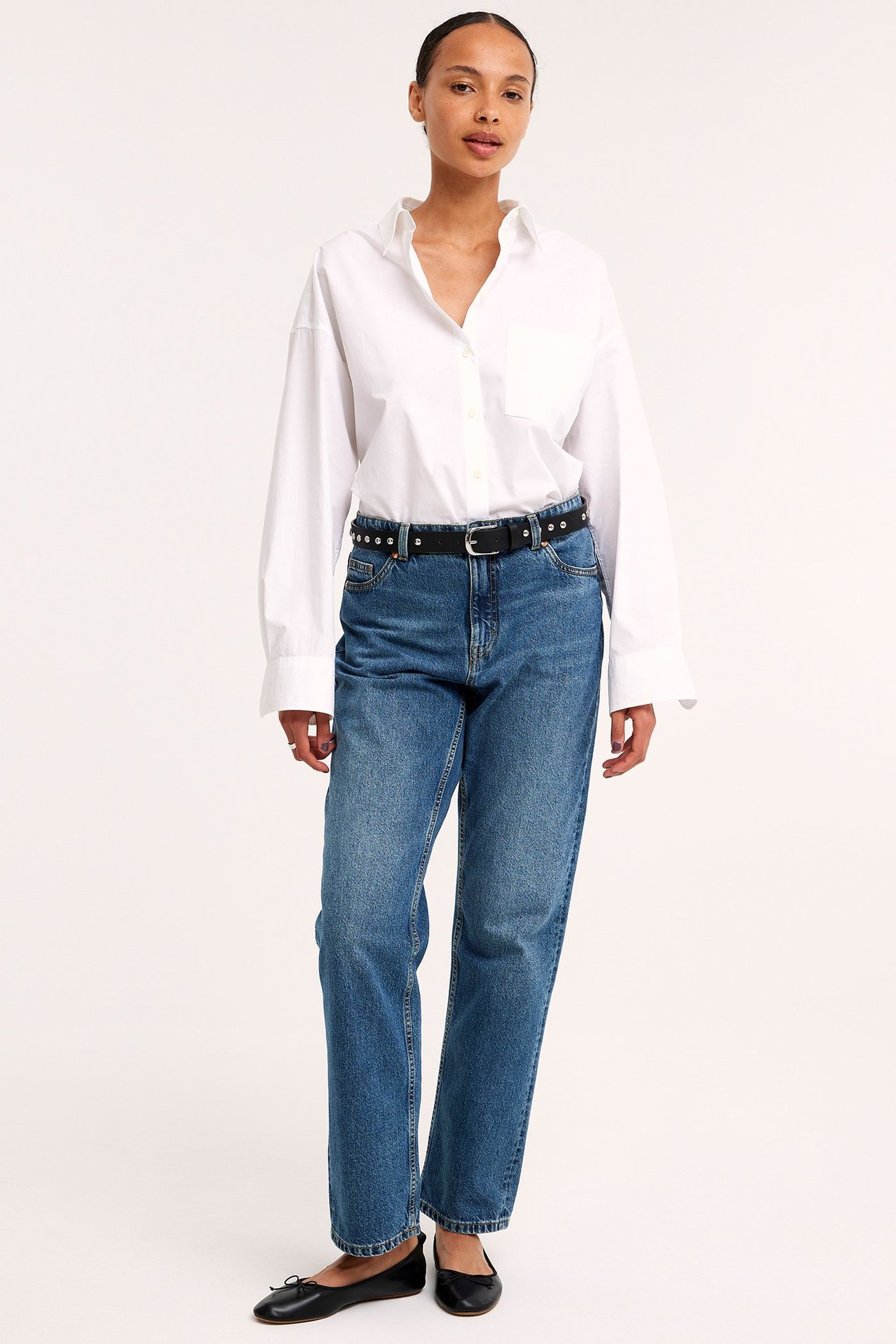 Framifrån: Kvinna i blå raka jeans, vit oversized skjorta, svart nitbälte och svarta ballerinaskor.