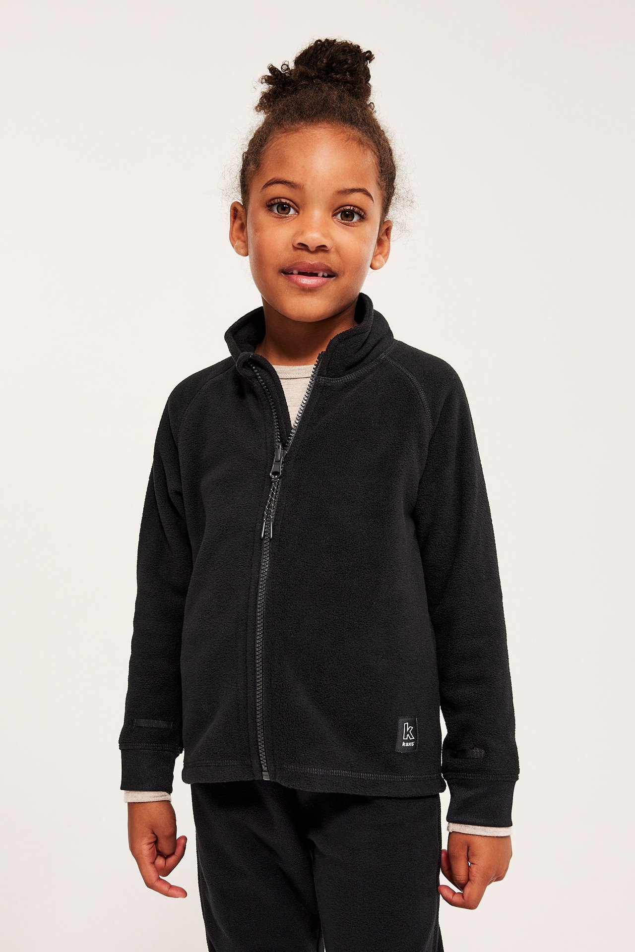 Edestä kuvattu lapsi mustassa vetoketjullisessa fleecetakissa, vaaleanbeigen paidan ja mustien housujen päällä.
