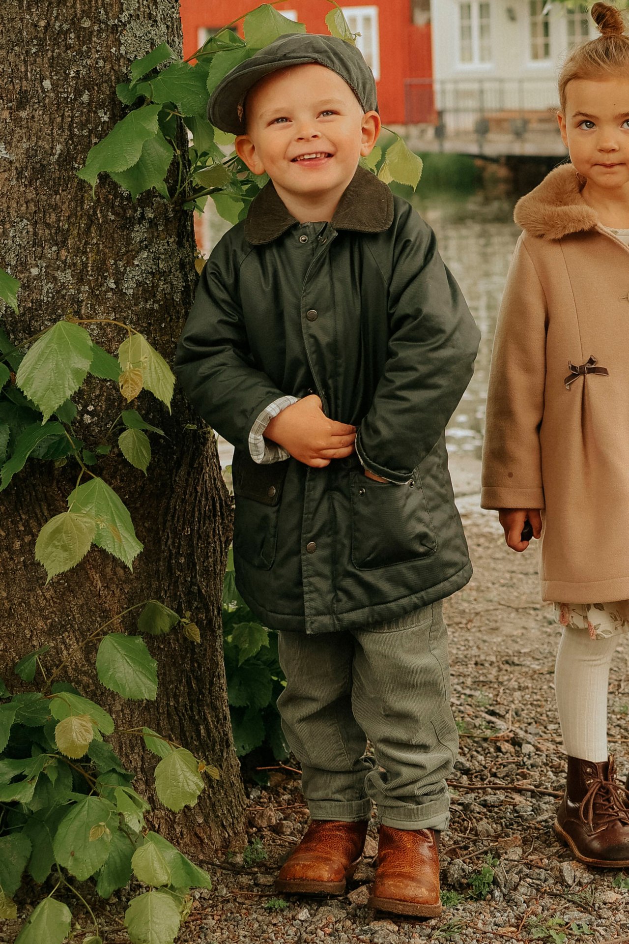Framifrån: Barn i grön vaxad jacka, mössa, byxor och bruna kängor. Flicka i beige kappa.