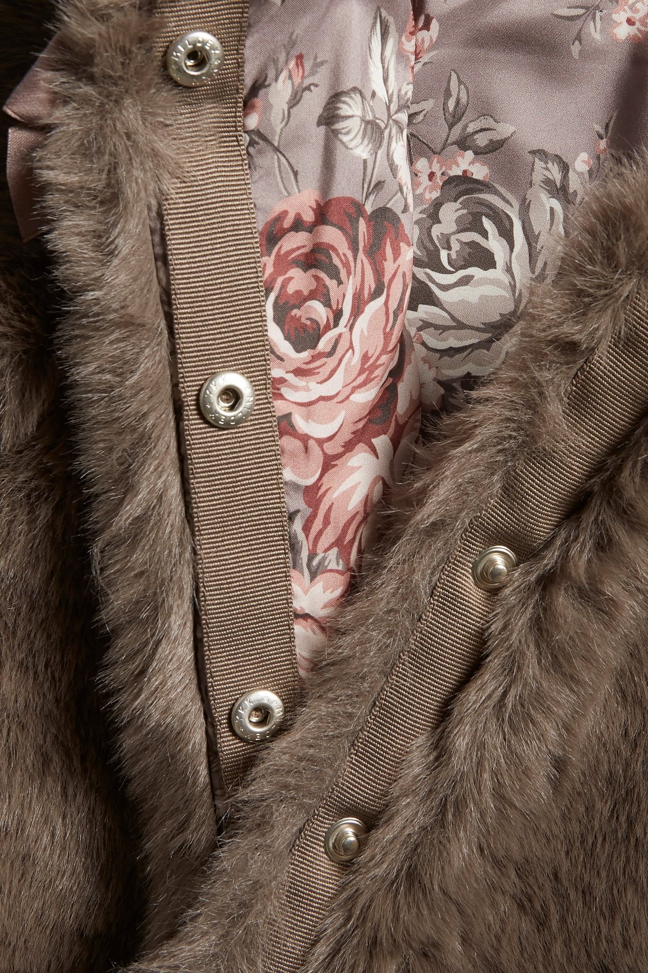 Nærbilde av en brun fuskepelsjakke med dekorativ sløyfe, skjulte trykknapper og blomstret fôr.