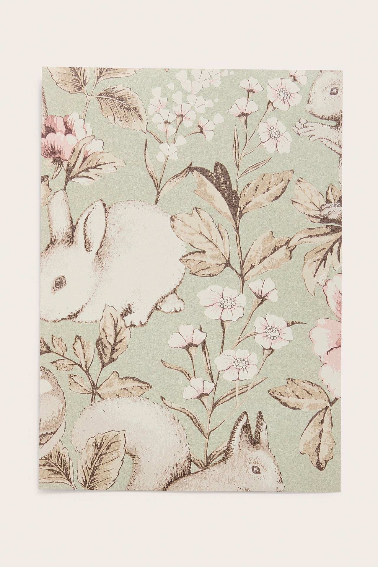 Närbild av ett grönt tapetprov med vita kaniner, ekorrar, rosa-vita blommor och bruna löv.