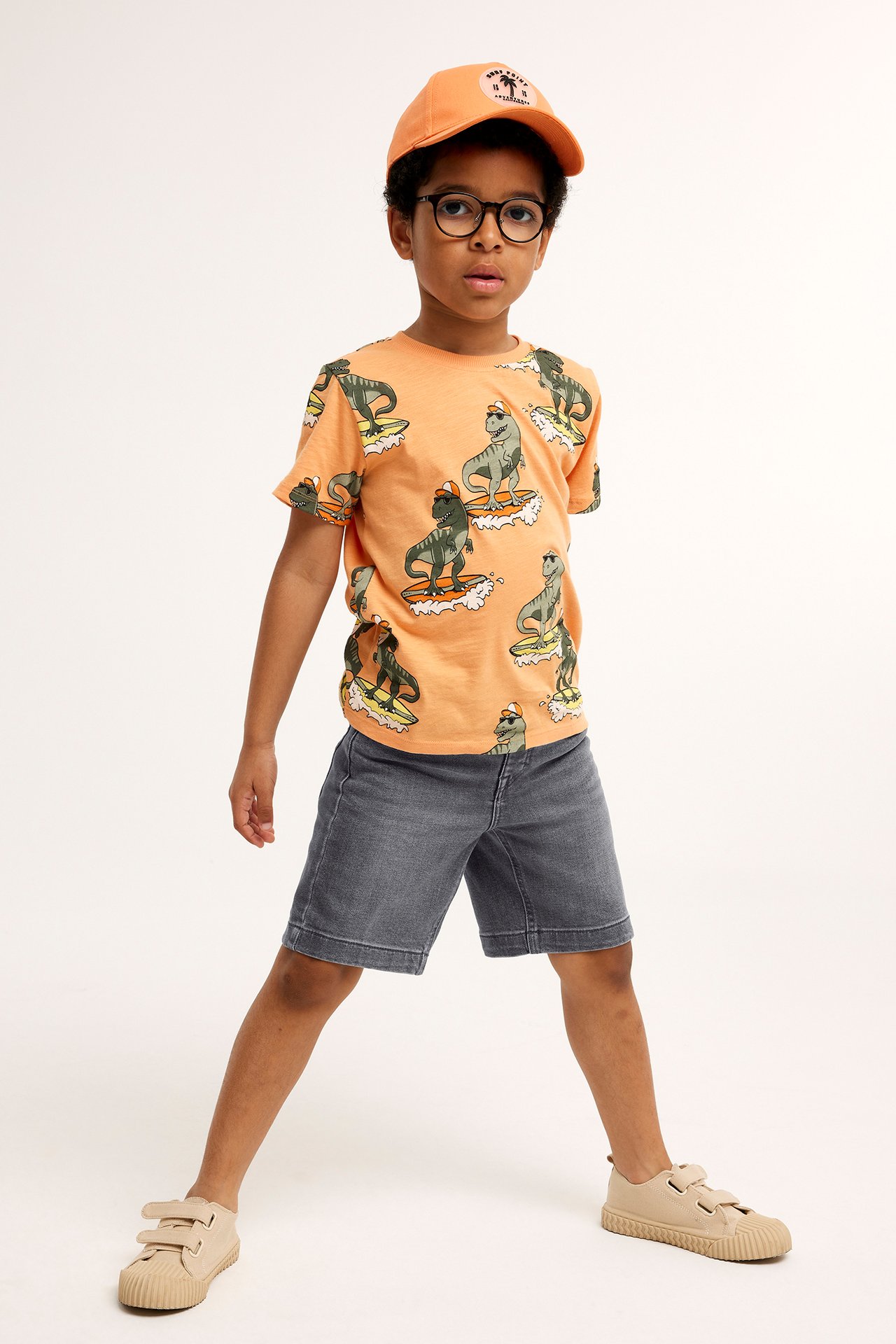 Framifrån: Barn i ljusgrå stretchjeansshorts, orange dinosaurie-t-shirt, keps, glasögon, sneakers.