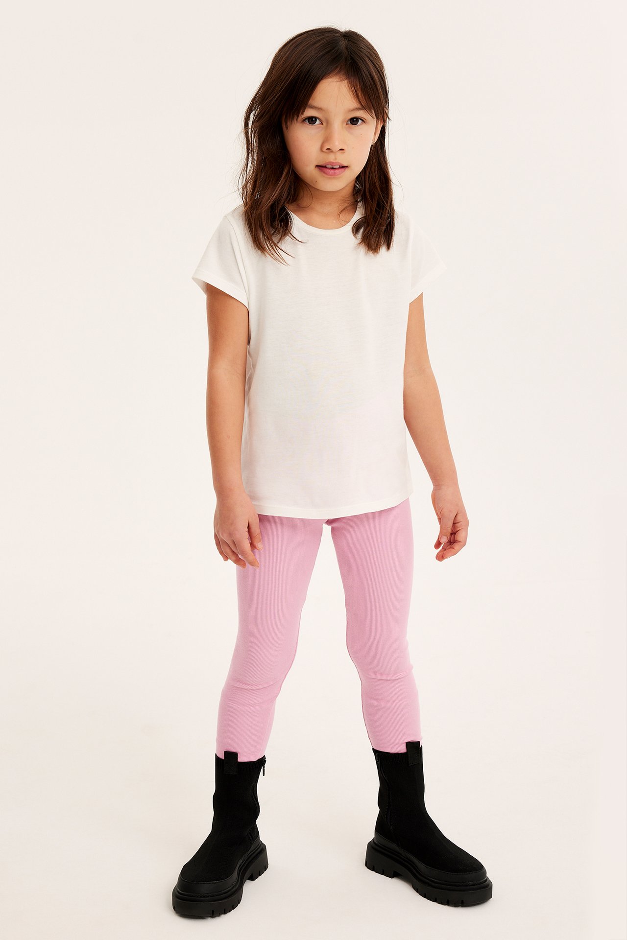 Frontvy av ett barn i rosa smala ribbade leggings, en vit t-shirt och svarta grova stövlar.