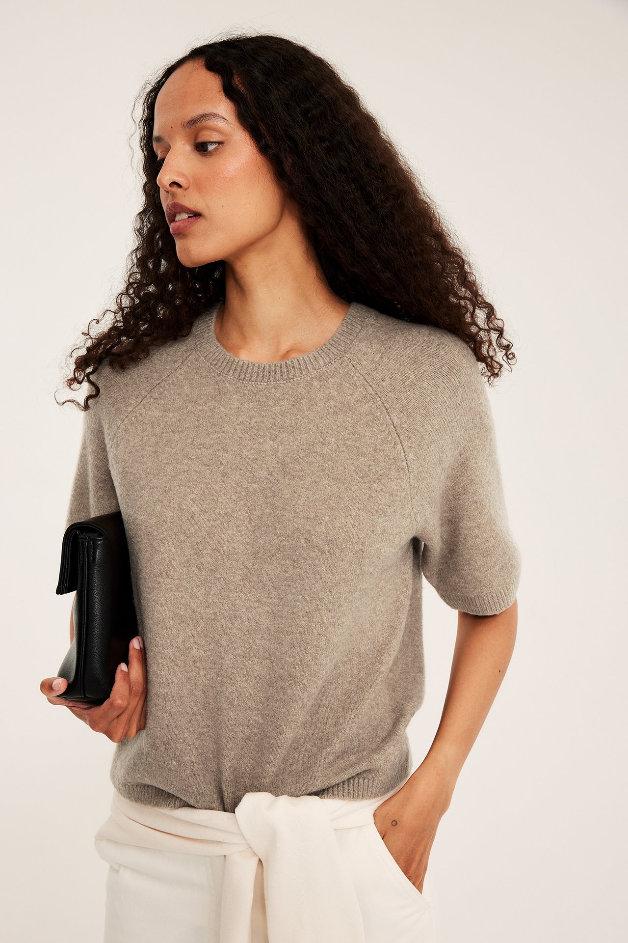 Kobieta w brązowym melanżowym dzianinowym T-shircie oversize, z czarną kopertówką i białymi spodniami.