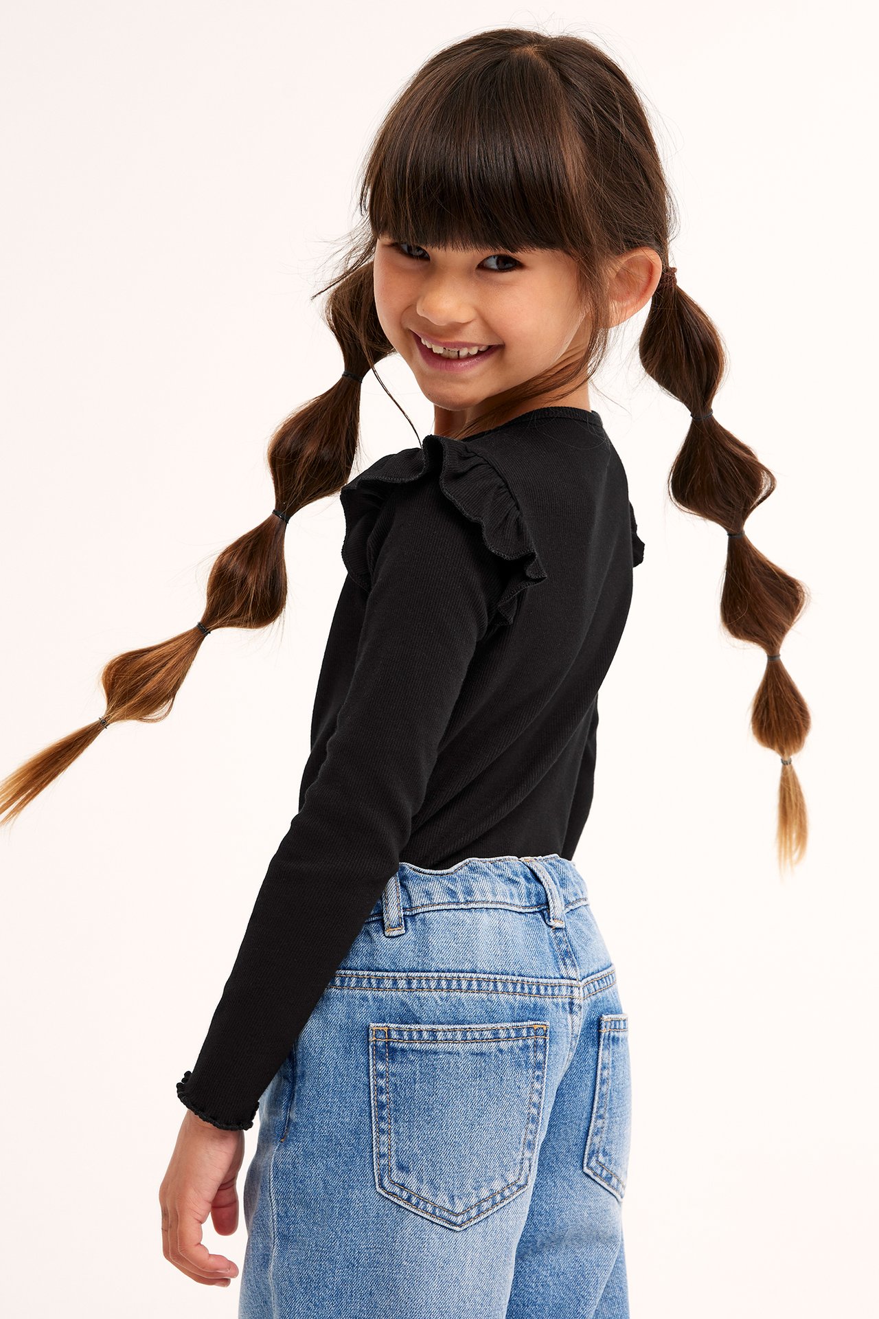 Bakifrån på ett barn i svart ribbad långärmad topp med volanger och blå jeans.