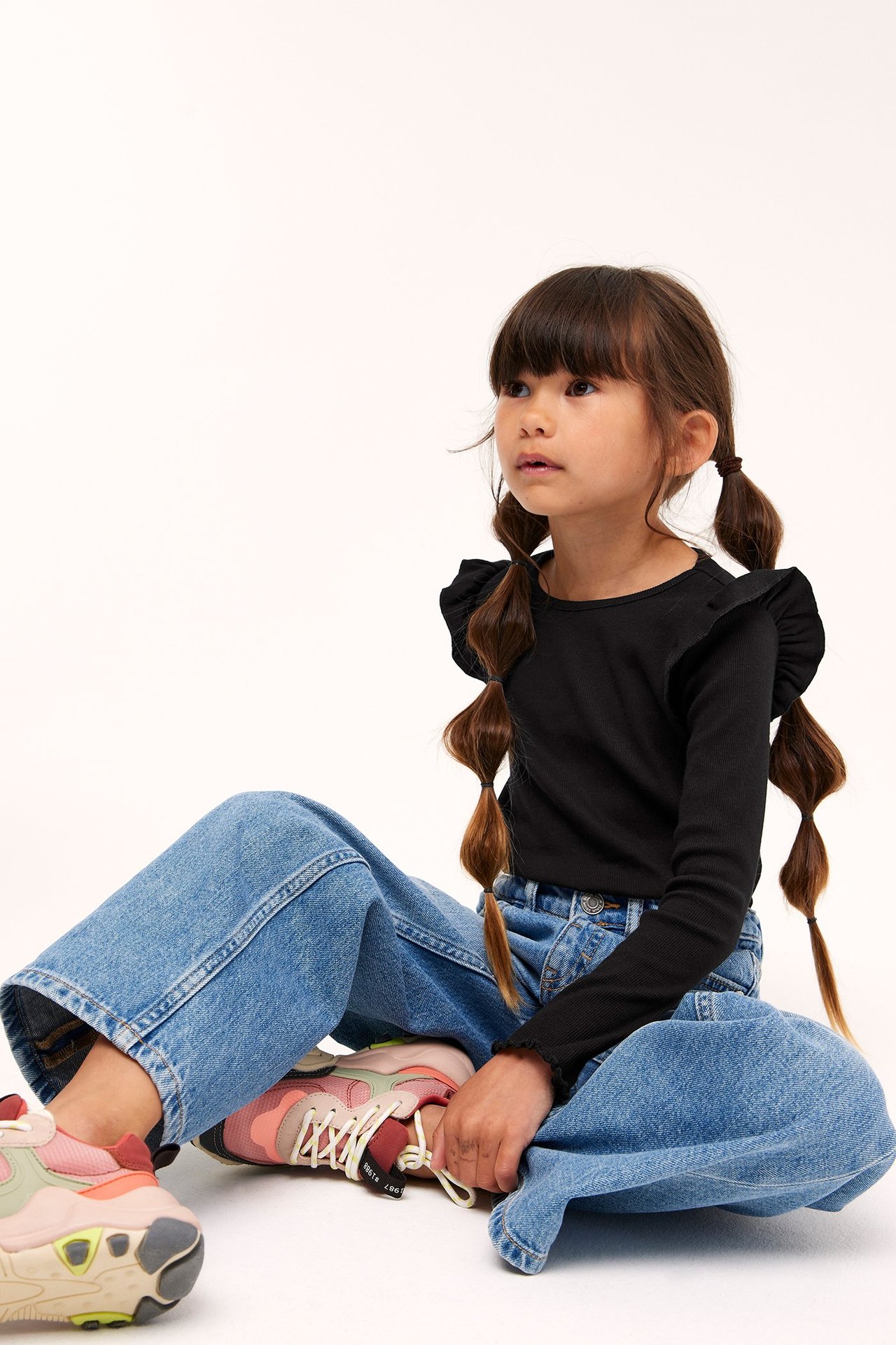 Edestäpäin: lapsi mustassa ribbineuleisessa pitkähihaisessa topissa röyhelöillä, sinisissä leveälahkeisissa farkuissa ja monivärisissä lenkkareissa.