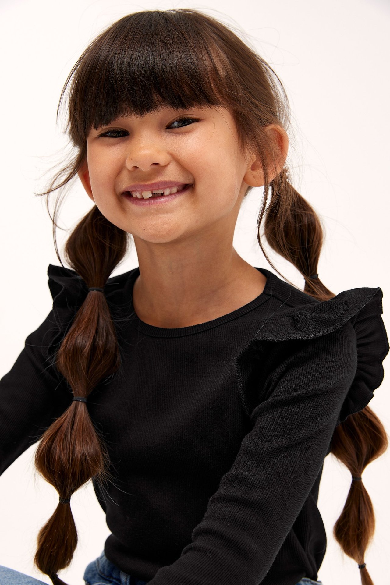 Lähikuva edestä lapsesta, jolla on musta ribattu pitkähihainen toppi röyhelöillä olkapäillä.