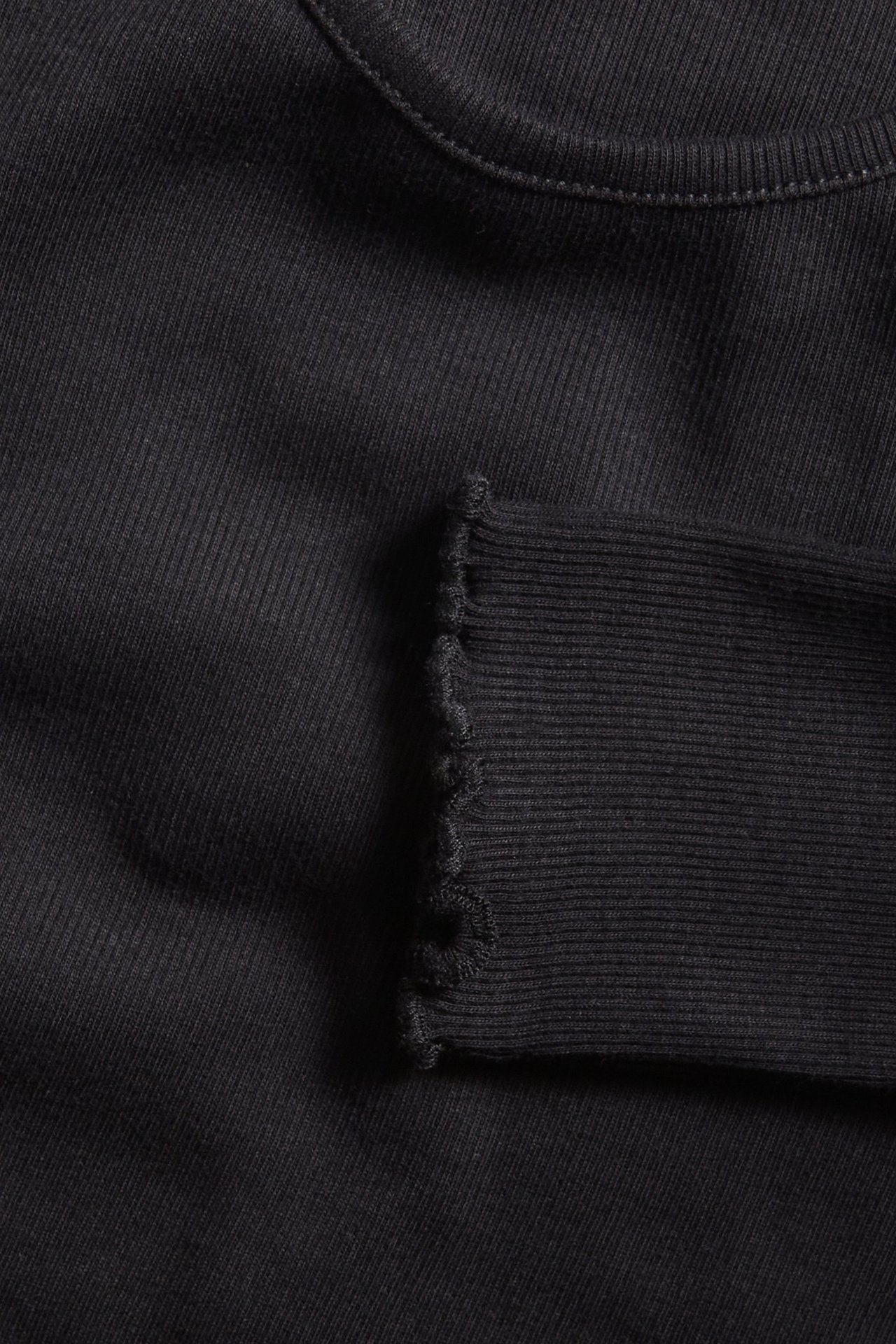 Zbliżenie czarnego prążkowanego topu z długim rękawem, z falbanką na mankiecie i okrągłym dekoltem.