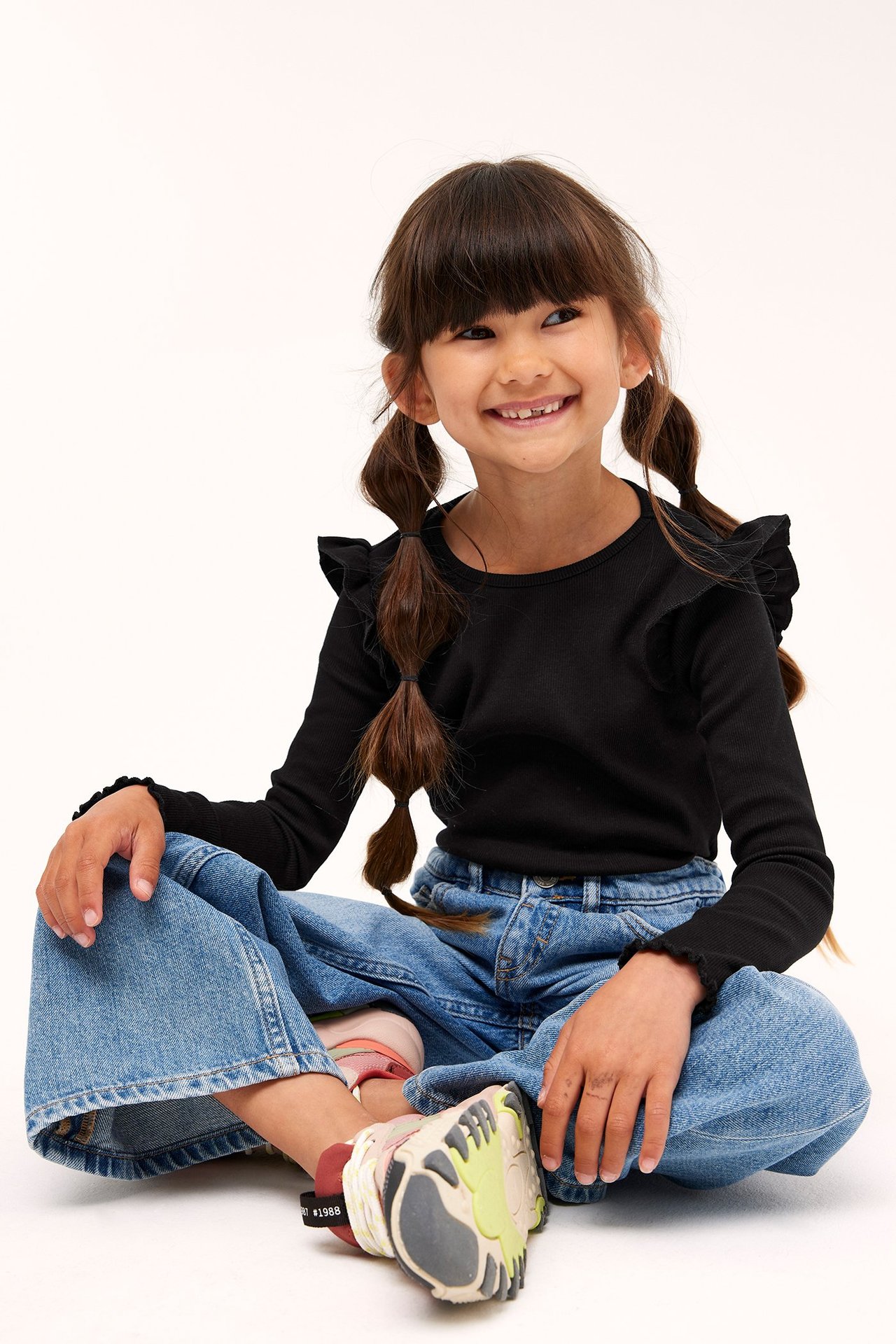 Widok z przodu: Dziecko w czarnym prążkowanym topie z falbankami, niebieskich dżinsach i sneakersach.