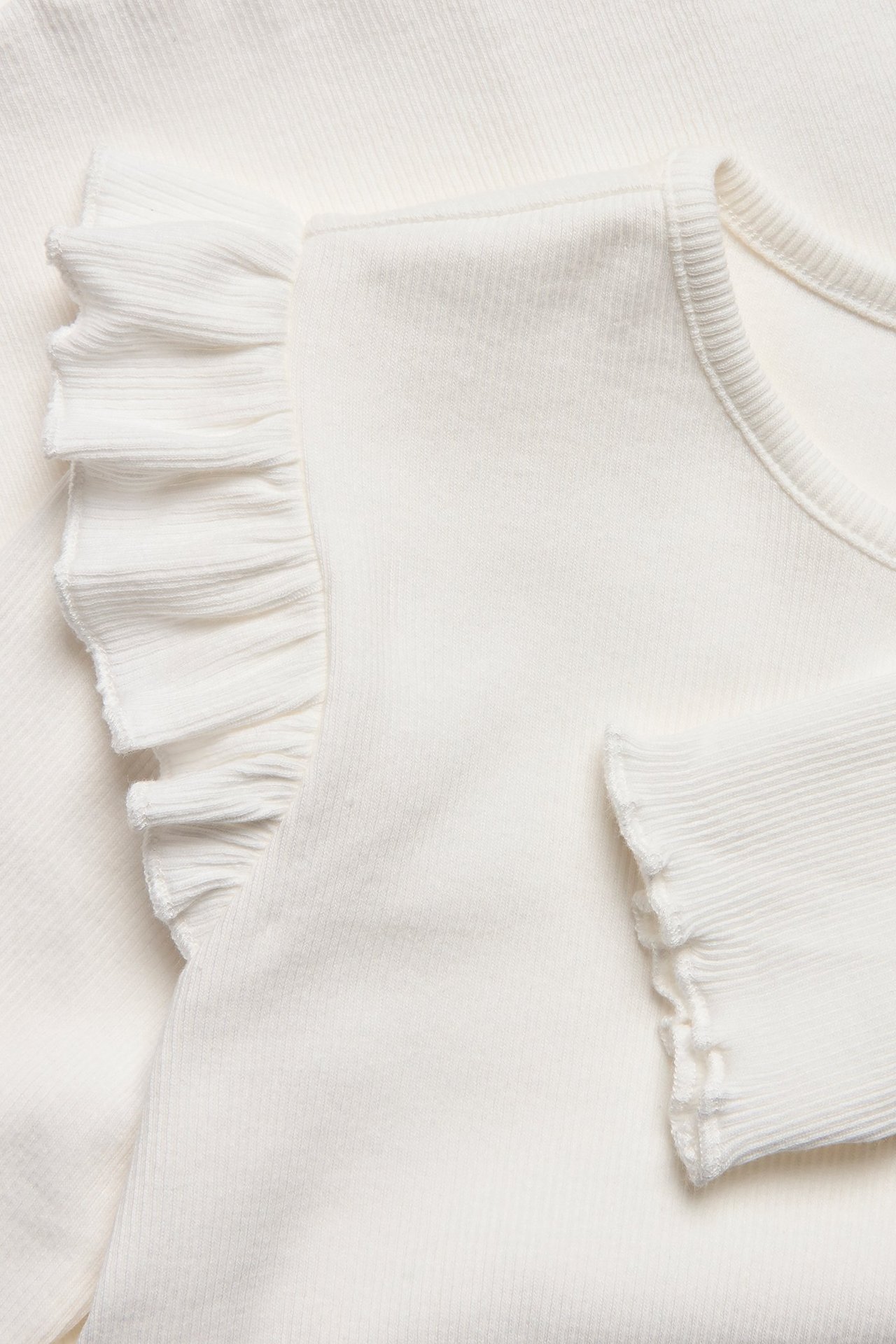 Zbliżenie białego prążkowanego topu z długim rękawem, z falbankami na ramieniu i mankietach.