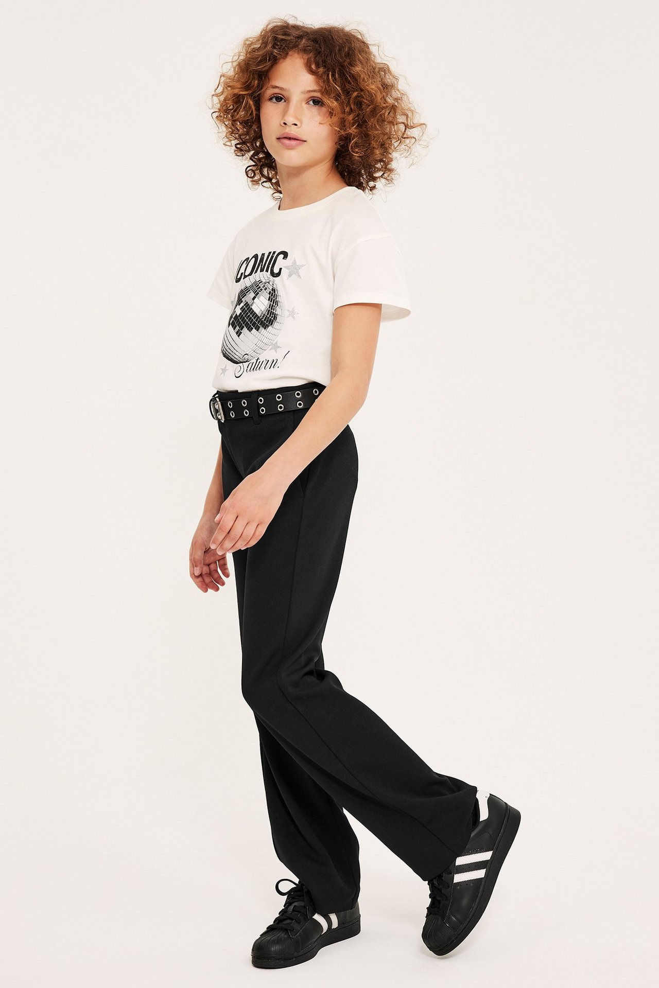 Sidebilde: Et barn i svarte jersey dressbukser, hvit grafisk t-skjorte, svart naglebelte og svarte joggesko.