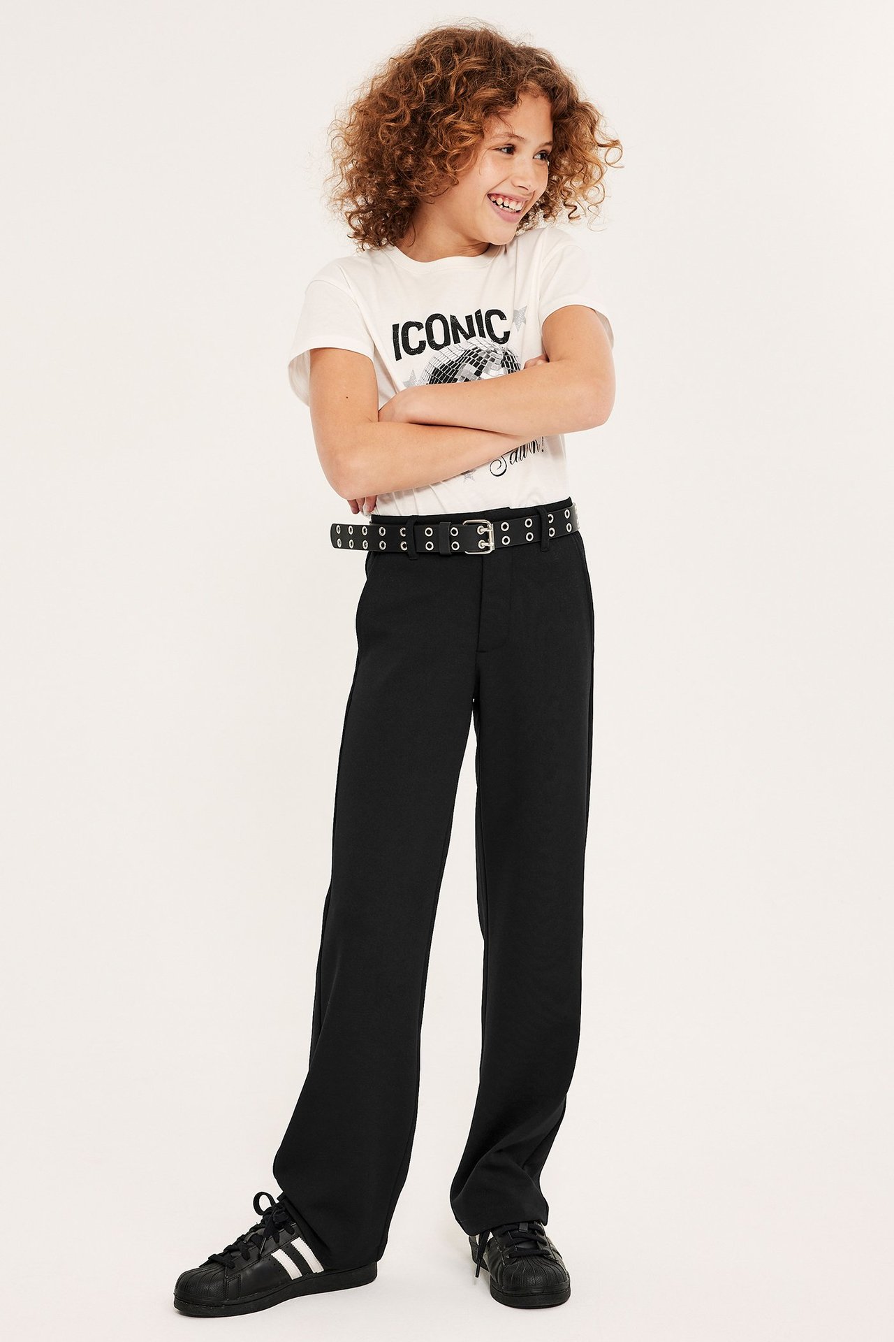 Widok z przodu dziecka w czarnych dzianinowych spodniach garniturowych, białej koszulce, czarnym pasku i czarnych sneakersach.