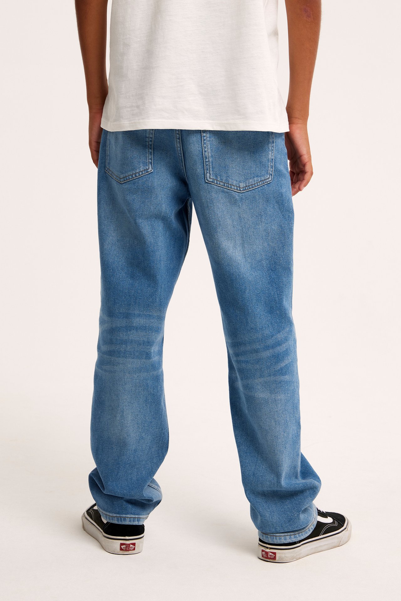 Widok z tyłu: Dziecko w jasnoniebieskich luźnych jeansach denim, białym t-shircie i czarnych trampkach.