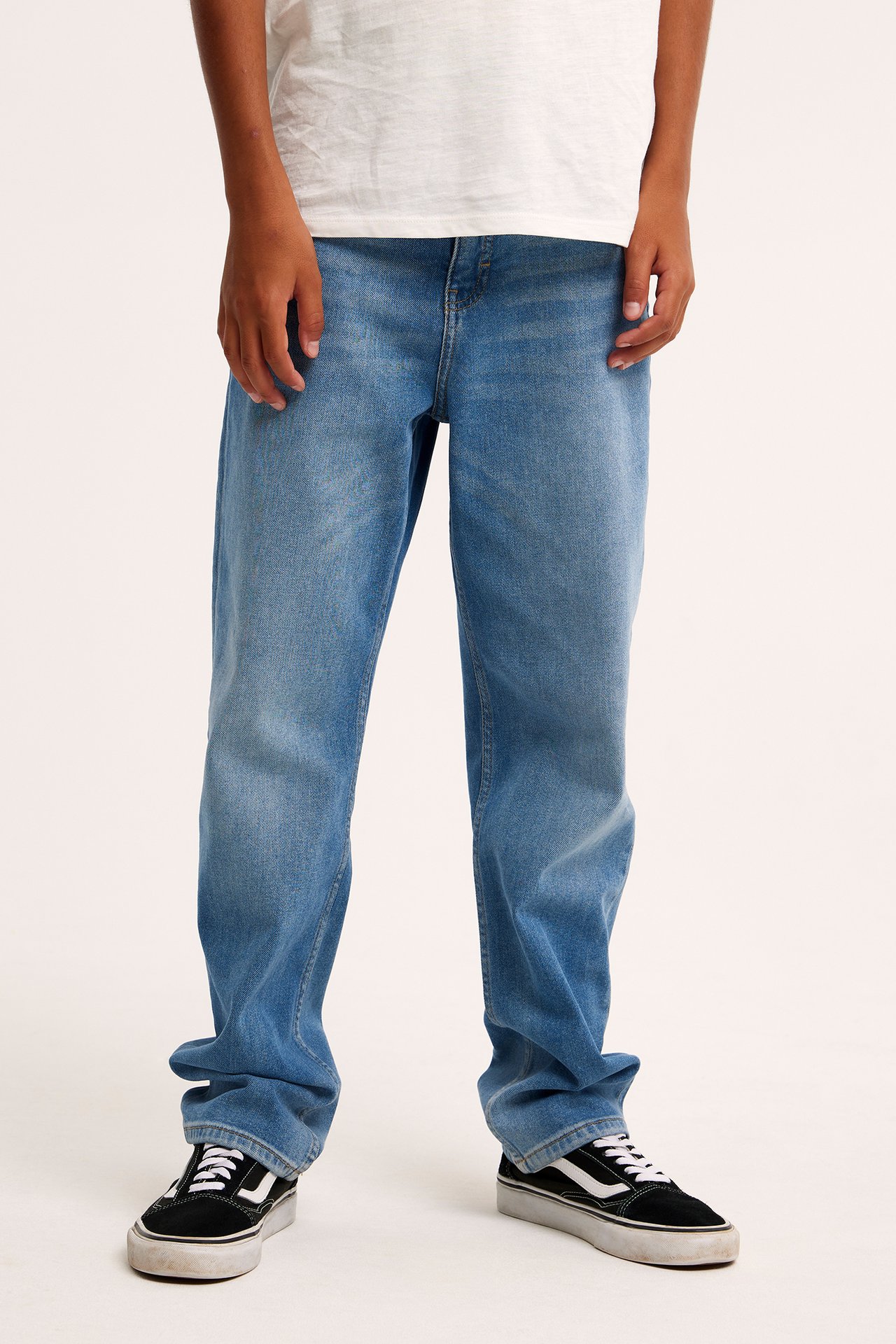Widok z przodu dziecka w jasnoniebieskich luźnych jeansach mid-waist, białym t-shircie i czarnych trampkach.