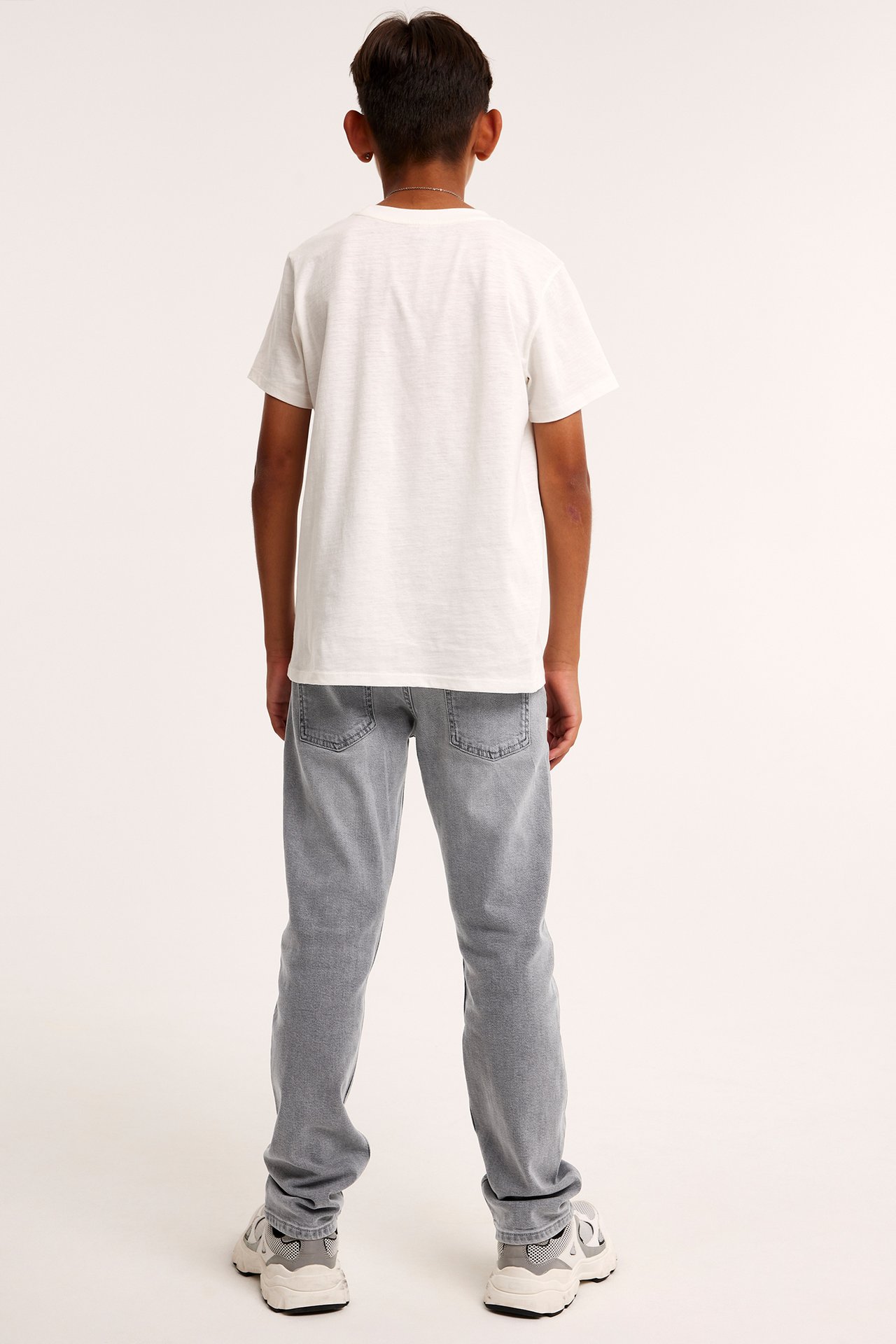Widok z tyłu dziecka w szarych prostych jeansach z wysokim stanem, białym T-shircie i białych sneakersach.