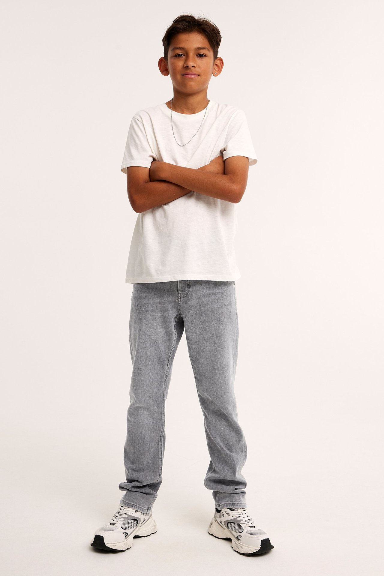 Widok z przodu dziecka w szarych prostych jeansach, białym t-shircie i białych sneakersach.