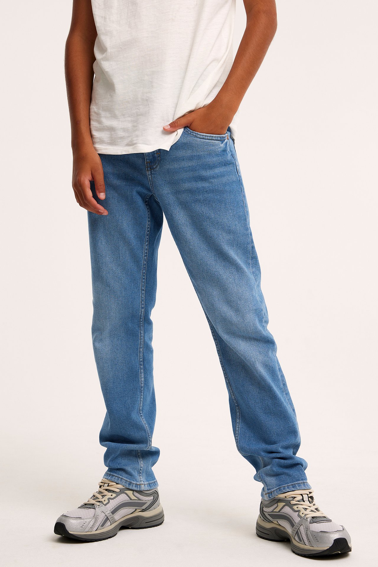 Widok z przodu dziecka w niebieskich prostych jeansach, białym T-shircie i szarych sneakersach.