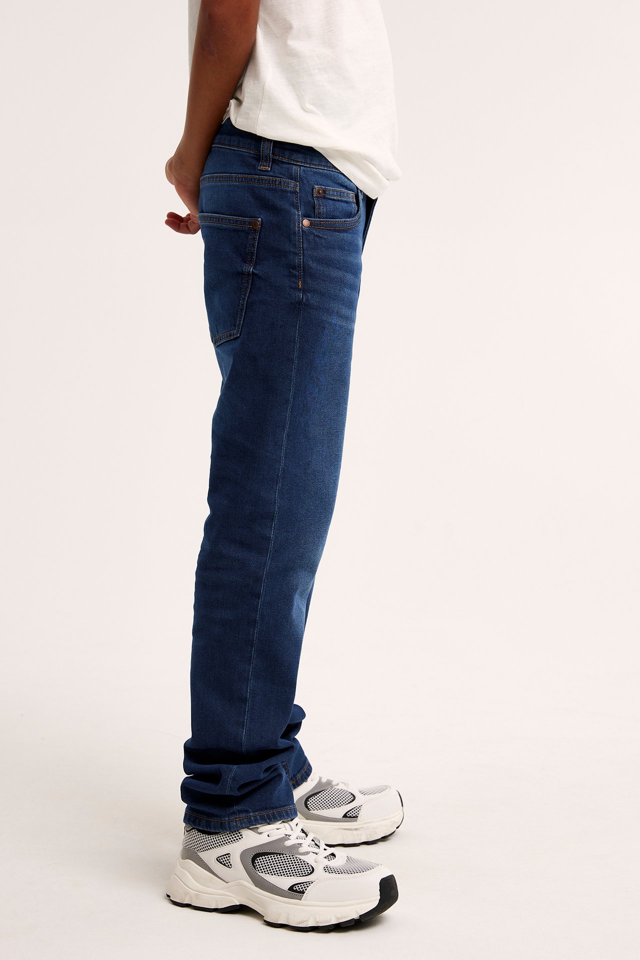 Widok z boku dziecka w ciemnoniebieskich prostych jeansach, białym t-shircie i biało-szarych sneakersach.