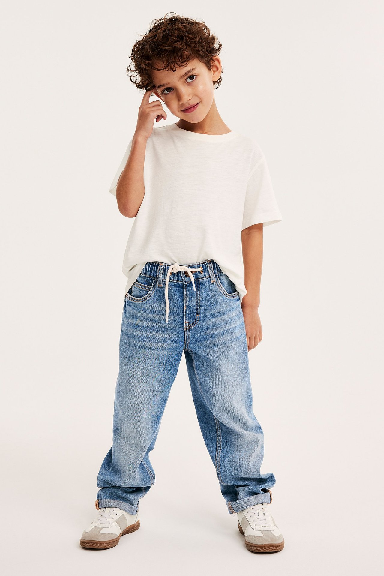 Widok z przodu dziecka w jasnoniebieskich jeansach relaxed, białym t-shircie i biało-beżowych sneakersach.