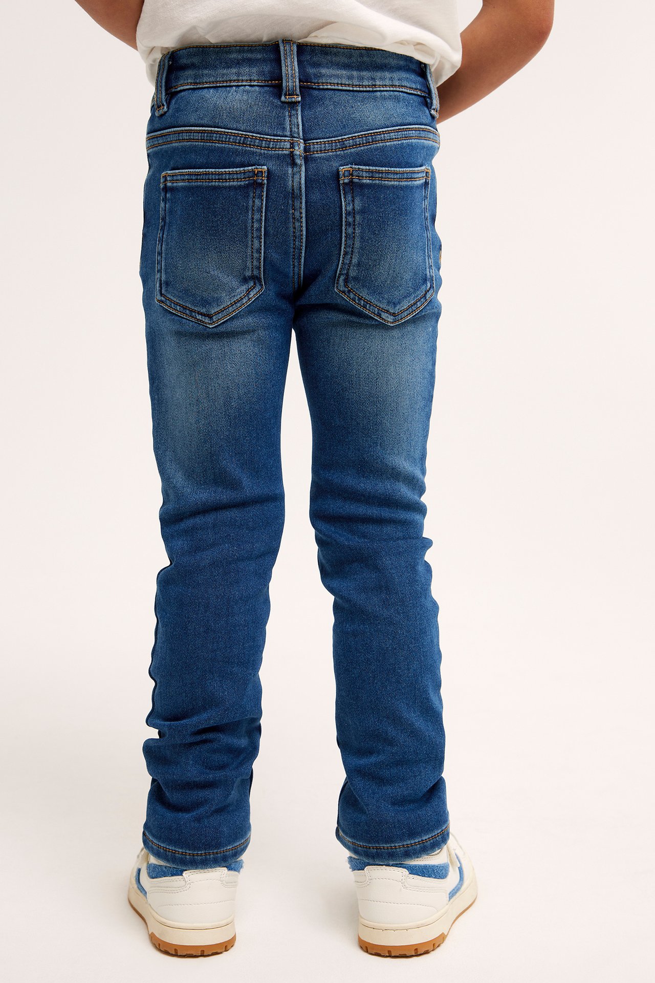 Bakfra av et barn i blå smale jogger-denimjeans, en hvit t-skjorte og hvite og blå joggesko.