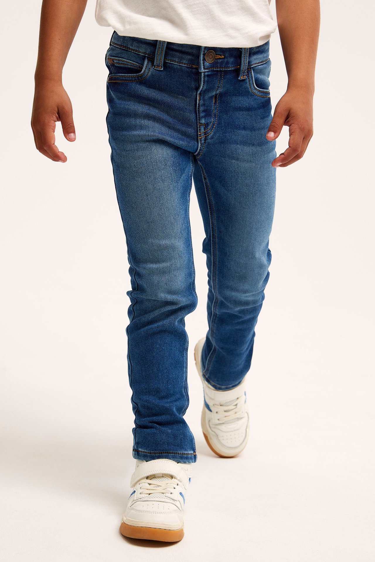 Edestä kuvattu lapsi sinisissä slim-fit jogger-farkuissa, valkoisessa t-paidassa ja valkoisissa lenkkareissa.