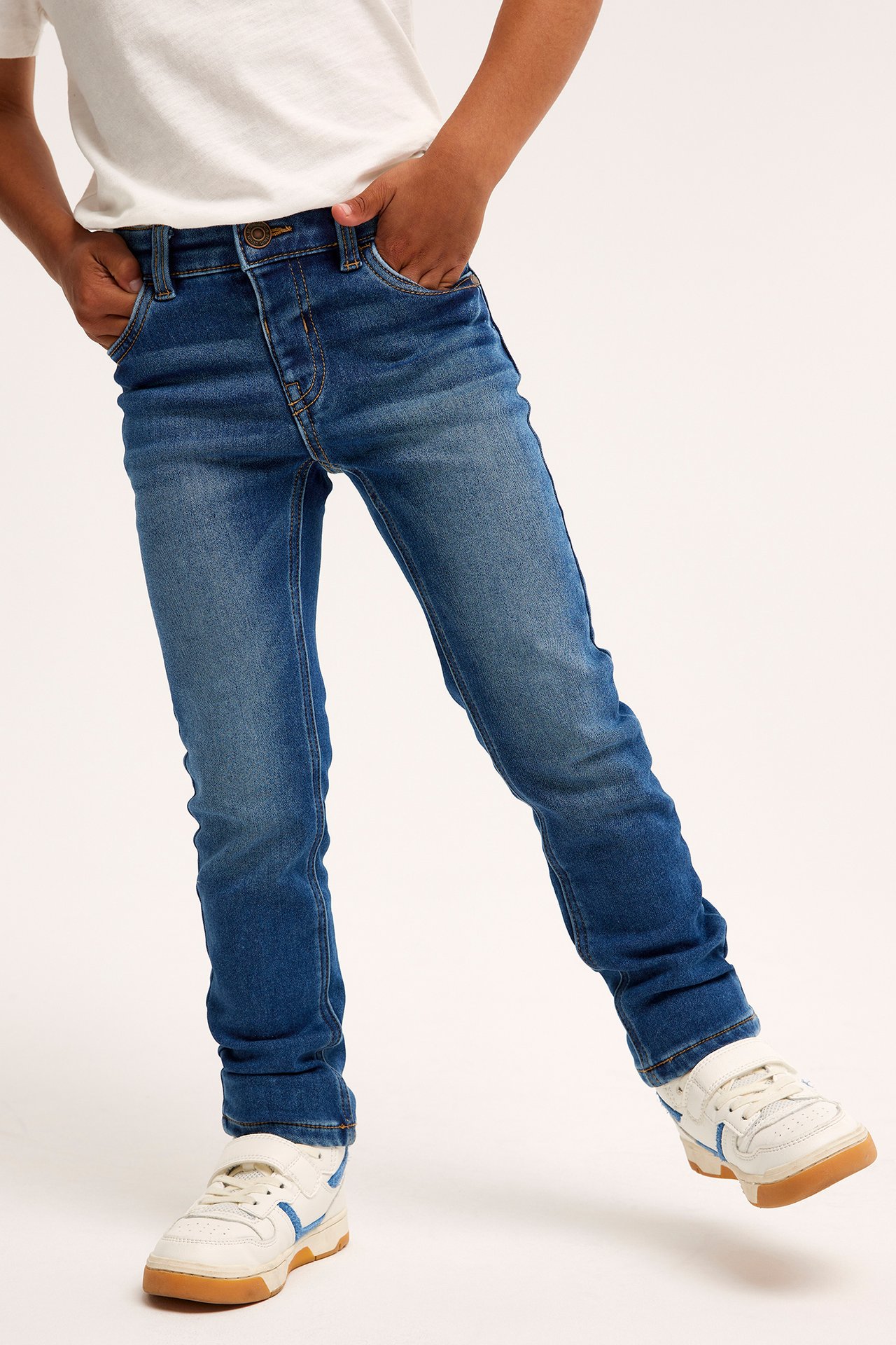 Forfra: et barn i blå slim-fit joggerdenim-jeans, en hvit t-skjorte og hvite joggesko.