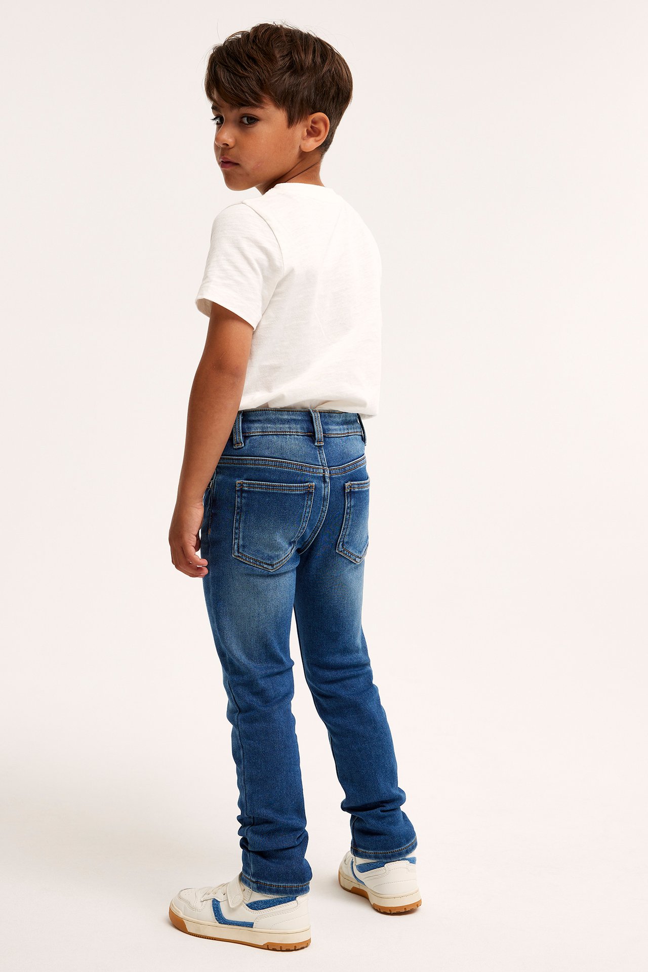 Bakfra: et barn i blå slim jogger denimjeans, en hvit t-skjorte og hvite joggesko.