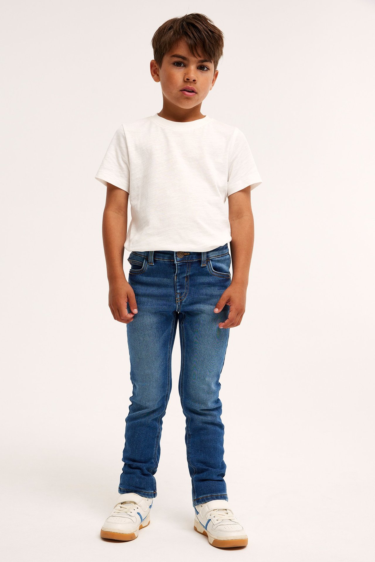 Lapsi edestä: siniset slim jogger denim -farkut, valkoinen t-paita ja valkoiset tennarit.
