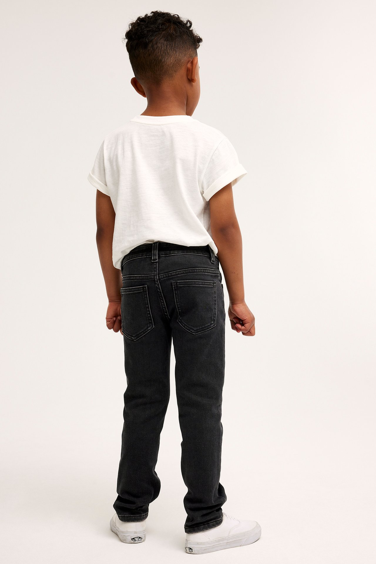 Lapsen takakuva mustissa slim jogger denimfarkuissa, valkoisessa t-paidassa ja valkoisissa lenkkareissa.