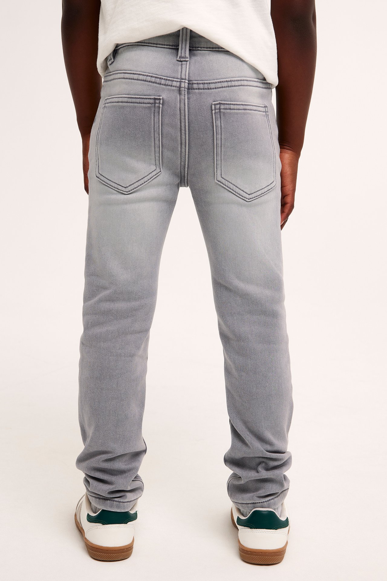 Bakifrån: ett barn i gråa slim-fit jogger denimjeans, en vit t-shirt och vita sneakers.